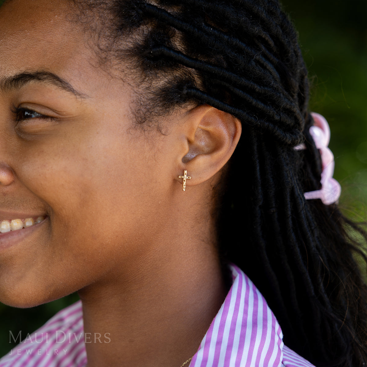 A side view of a smiling woman in a pink and white striped top wearing a Hawaiian Heirloom Old English Scroll Cross Earring in 14k yellow gold, against a blurred green background