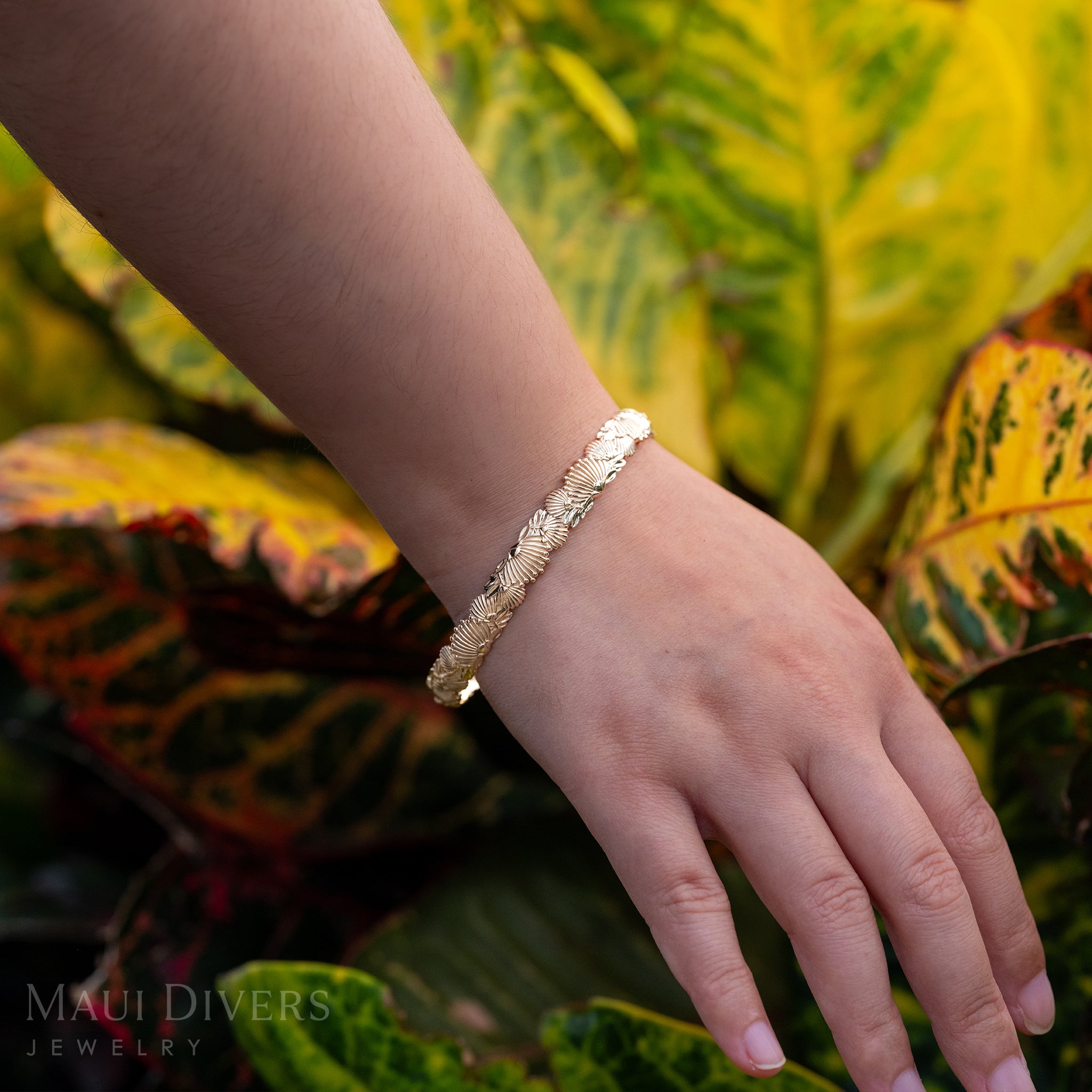 An ʻŌhiʻa Lehua Flower Patterned Bracelet in 14k yellow gold on a wrist, against a blurred yellow, red, and green leafy background