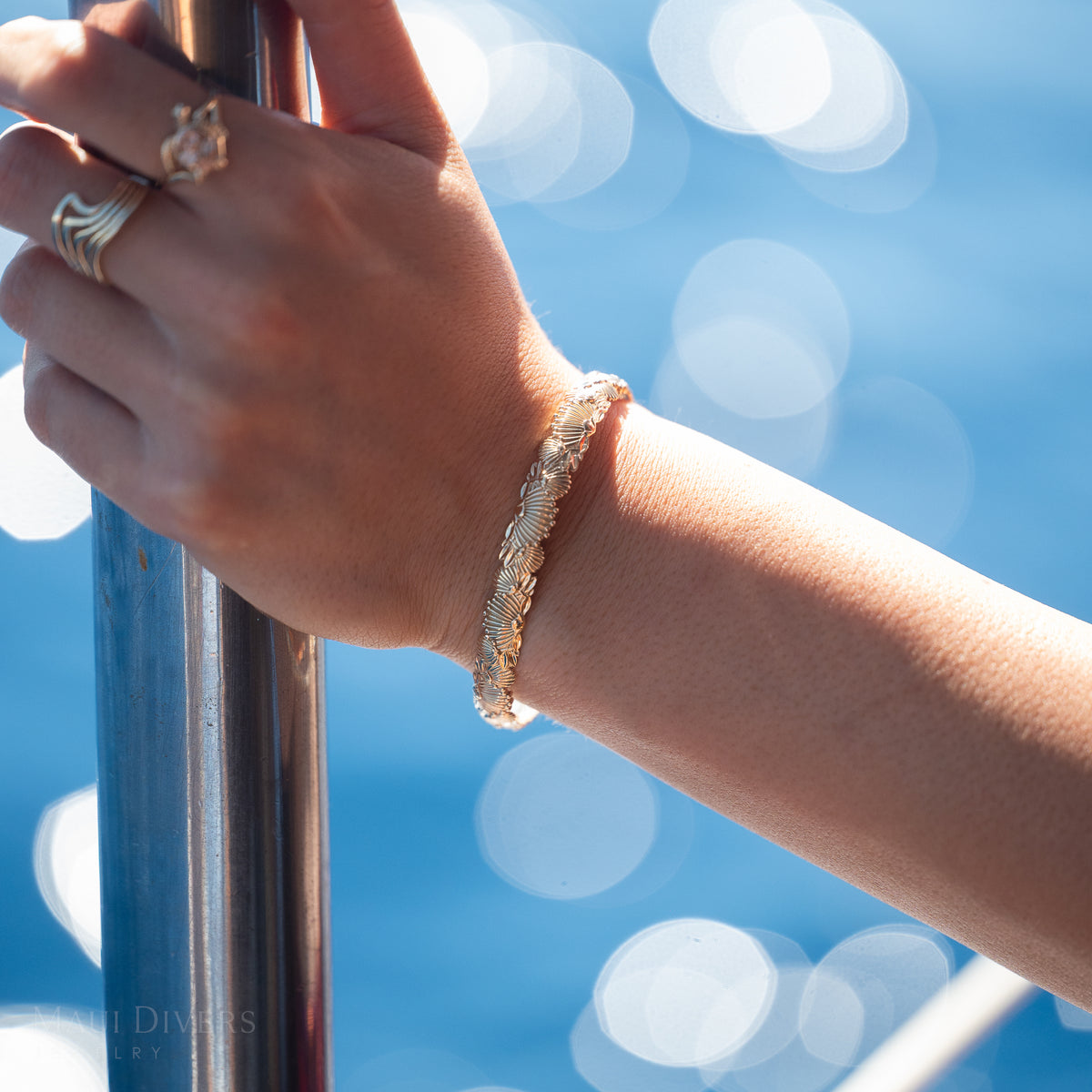 Close-up of a wrist wearing a gold ʻŌhiʻa Leahu patterned bangle against a blurry ocean background