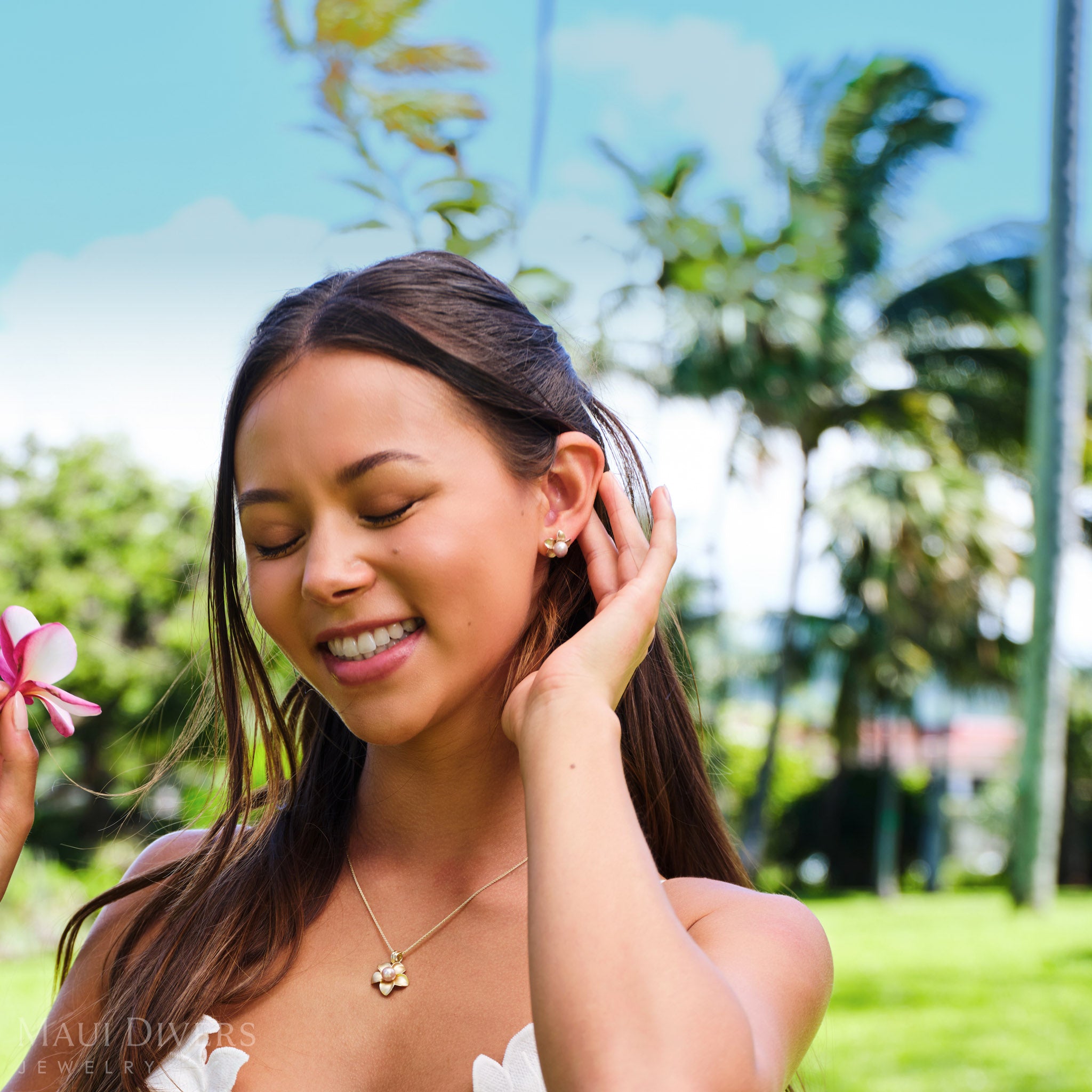 Smiling woman in a white top wearing a Pick A Pearl Plumeria Earring in 14k yellow gold with a pink pearl on her left ear, against a blurred park background