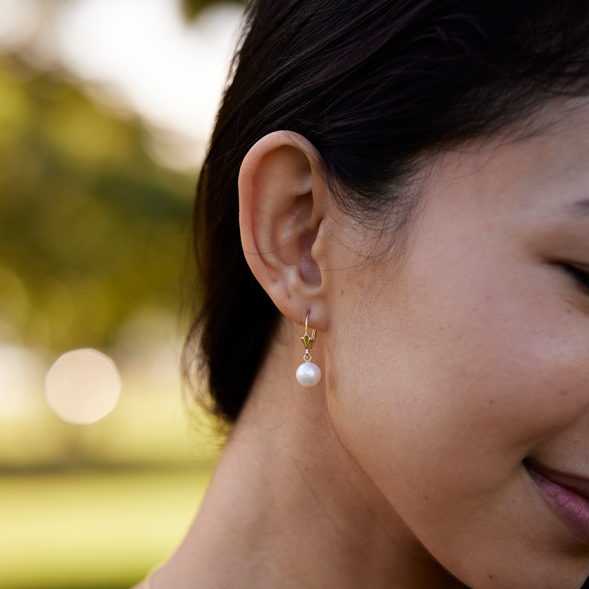 Pick A Pearl Earrings in Gold with Lavender Pearls on a woman's right ear with blurred tree background