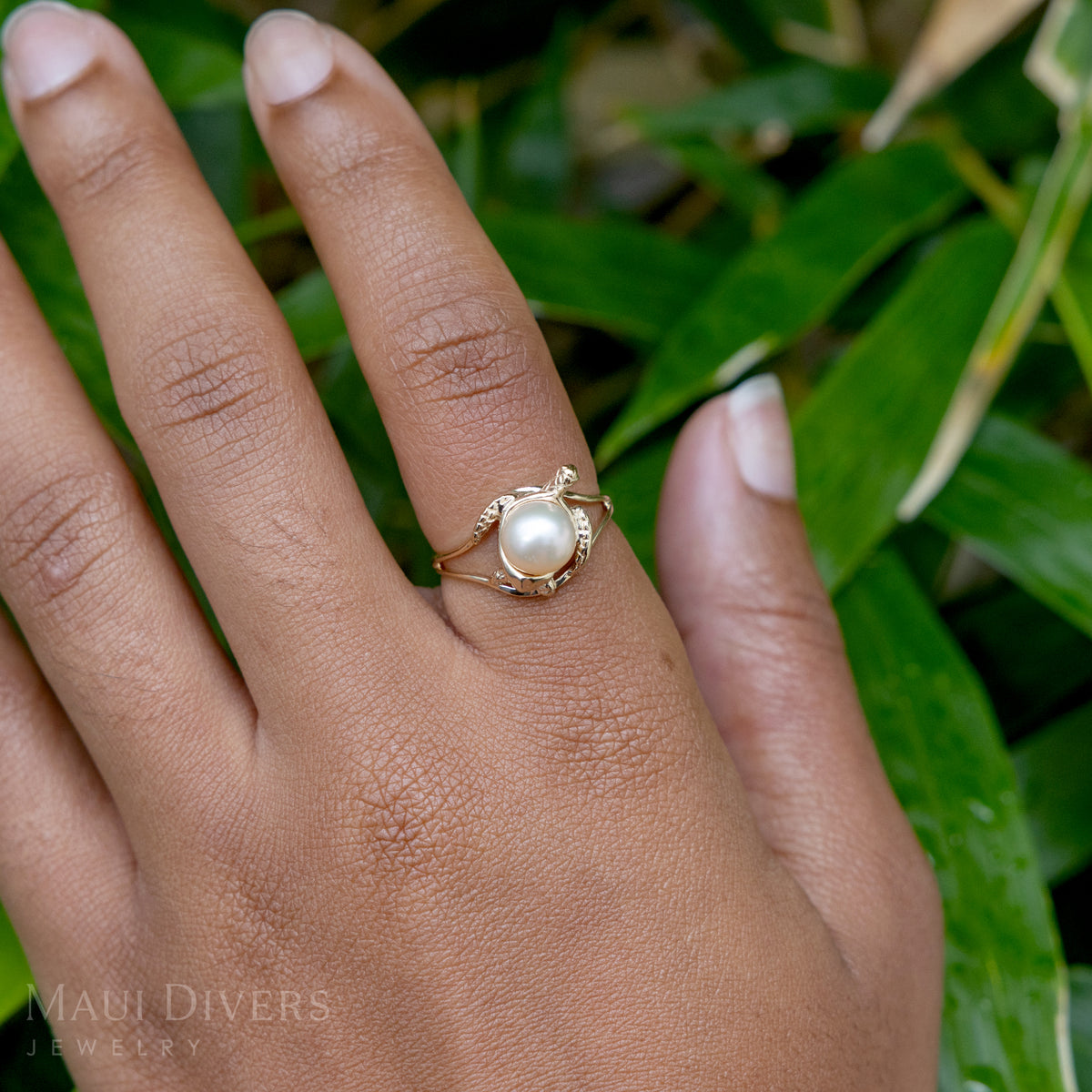 Close-up of a hand wearing a Pick A Pearl Honu Ring in 14k yellow gold with a freshwater white pearl, against a blurred leafy green background