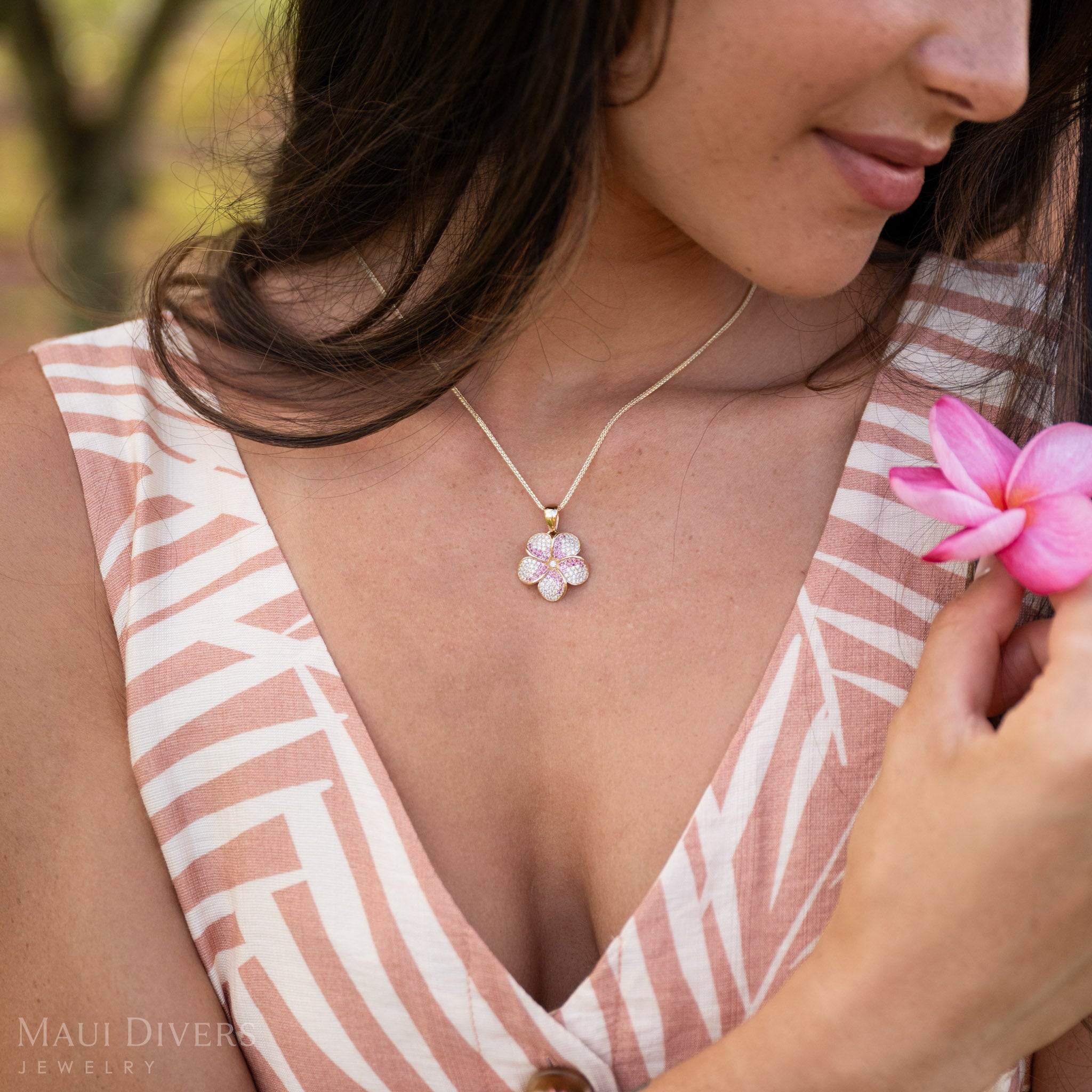 Woman wearing Plumeria Pink Sapphire Pendant in 14k Gold with Natural Diamonds and holding a pink flower to her heart.