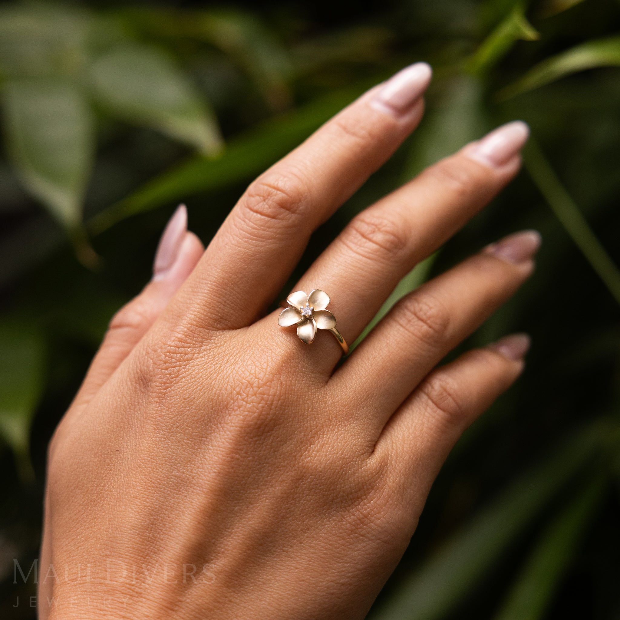 Hand wearing a Plumeria Ring in 14k yellow gold with a diamond against a blurred leafy background