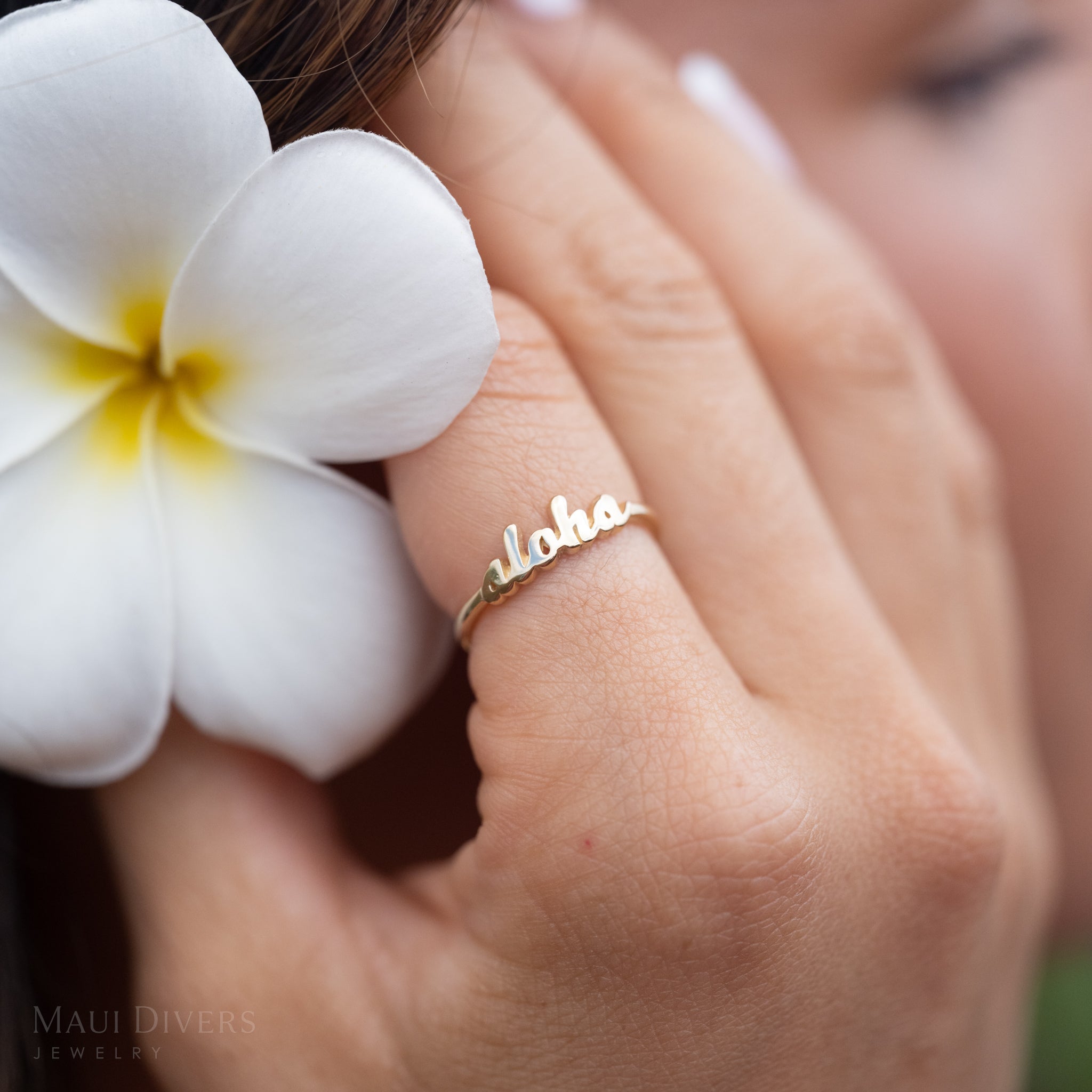 Hand wearing an ʻalohaʻ script ring in high-polish 14k yellow gold with a yellow and white plumeria in their hand