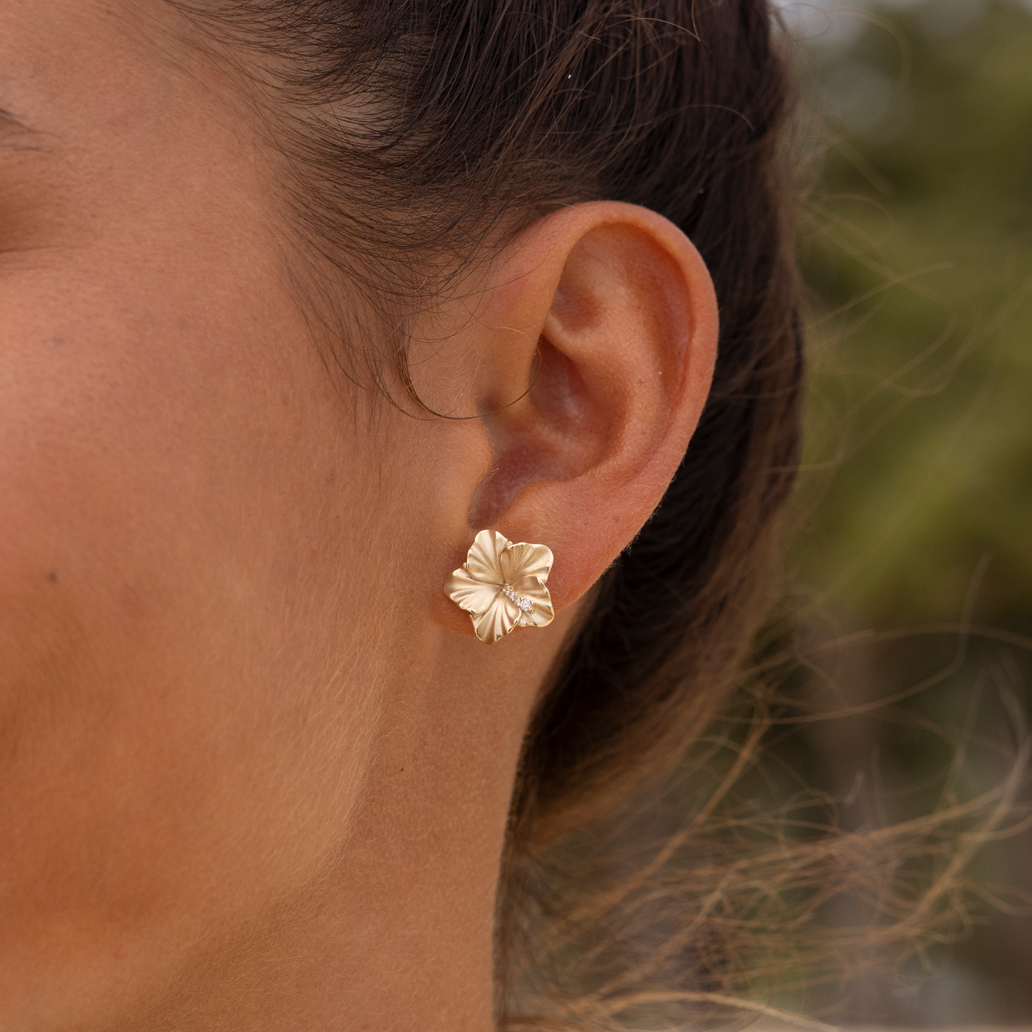 Close up of 14mm Diamond Hibiscus Stud Earrings in 14k Yellow Gold on ear with blurred nature background