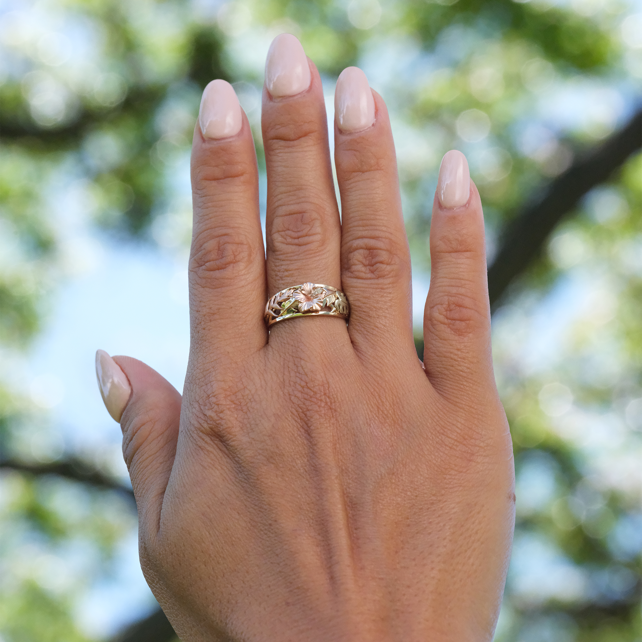 A woman's hand wearing a Hawaiian Gardens Hibiscus Ring in Tri Color Gold with Diamonds up in air against trees
