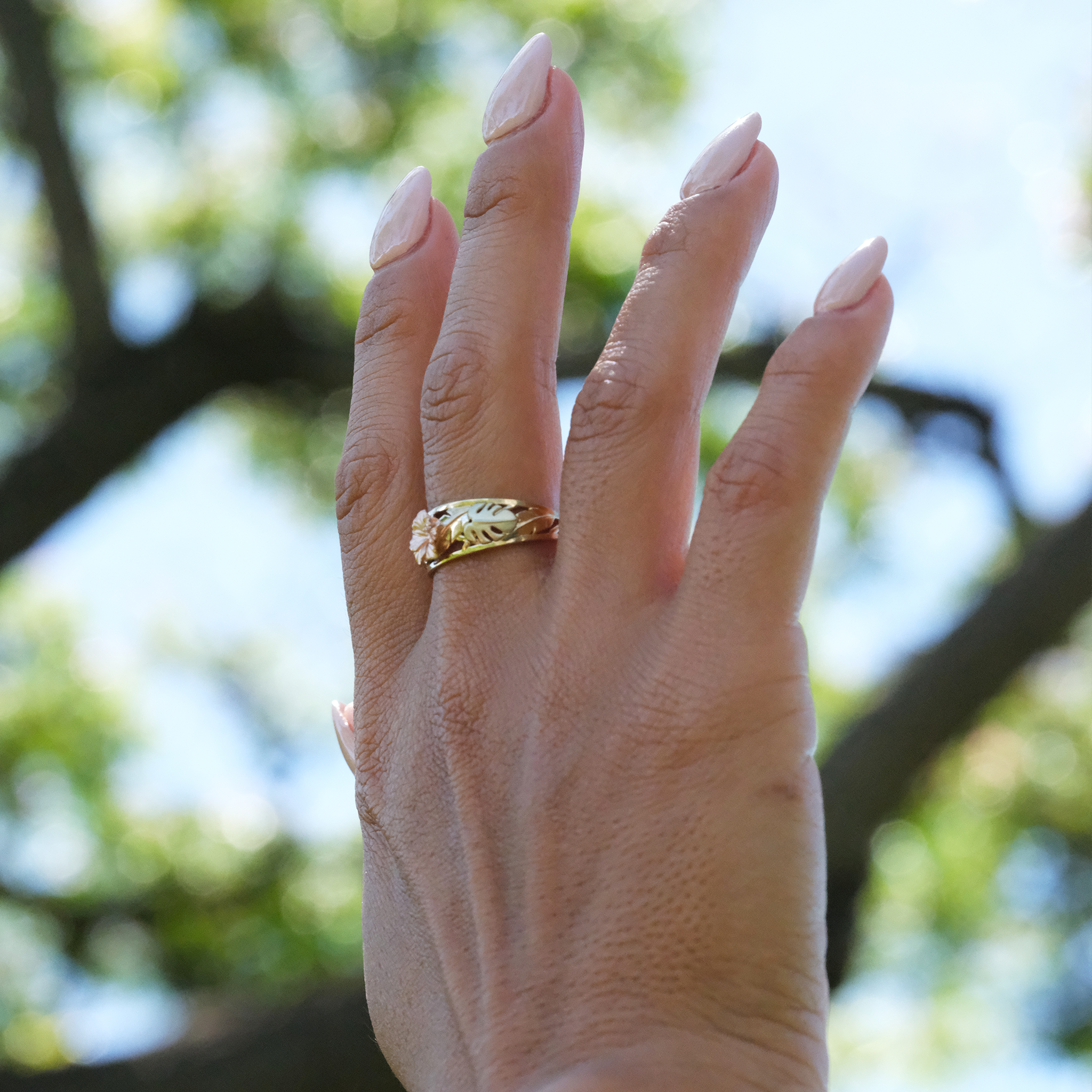 A woman's hand wearing a Hawaiian Gardens Hibiscus Ring in Tri Color Gold with Diamonds in the air against trees