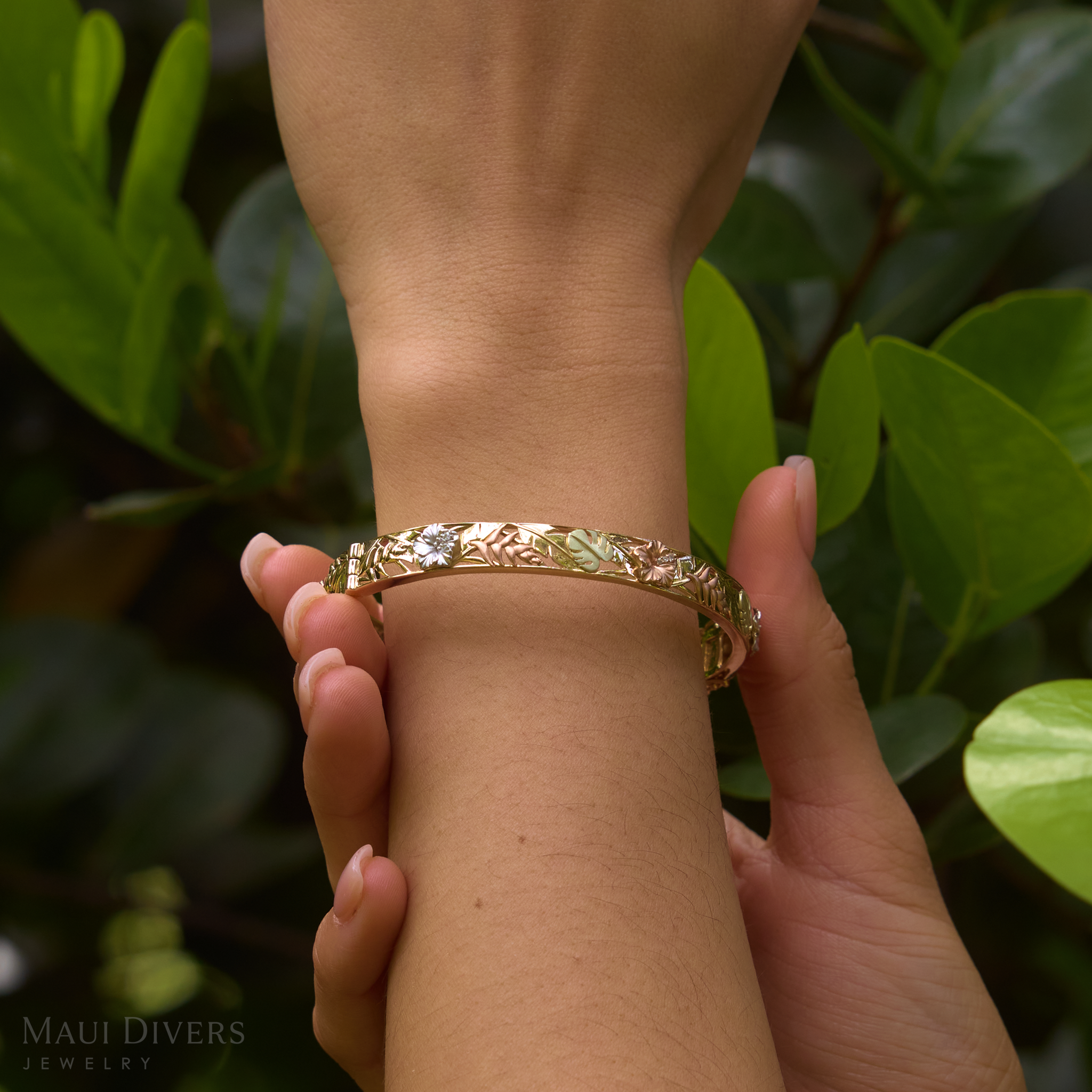 Hand wearing a Hawaiian Gardens Hinge Bracelet in Four Color Gold with Diamonds against green leaves