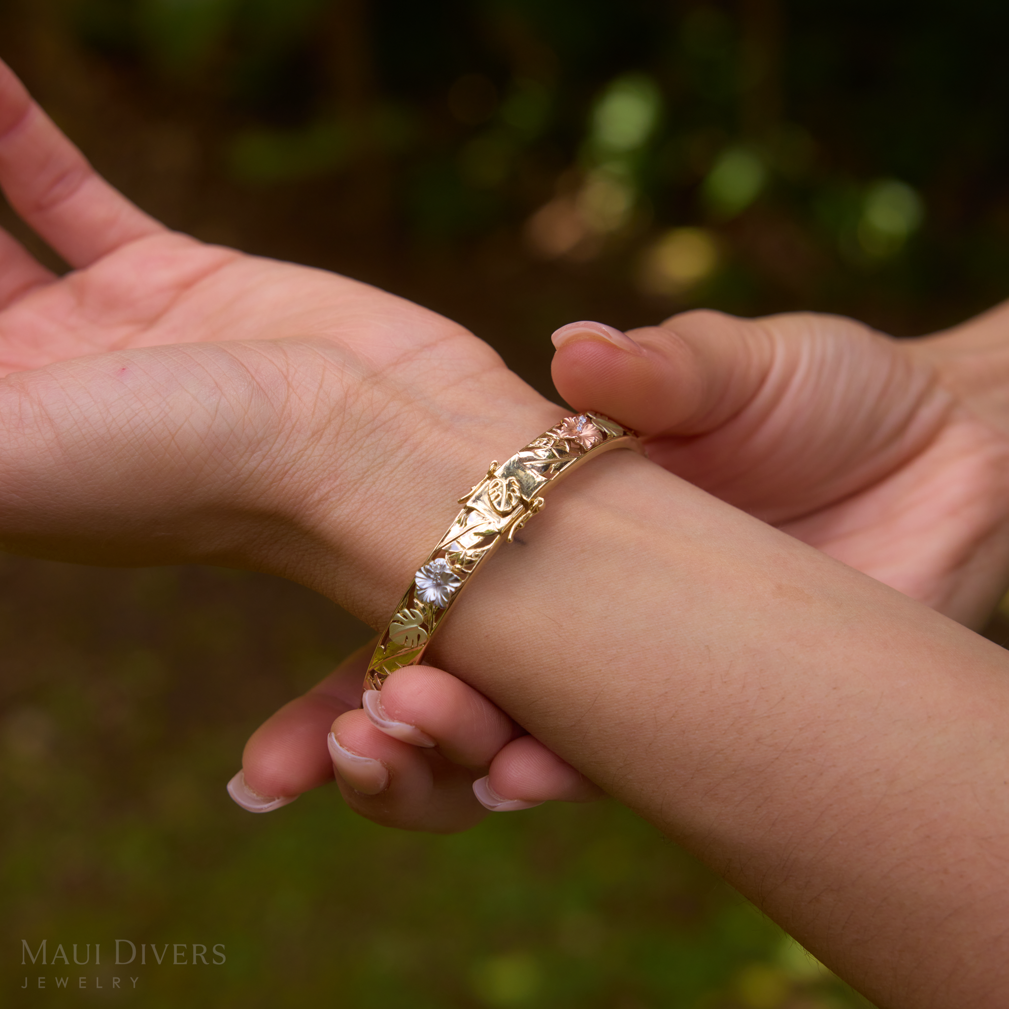 Hand wearing a Hawaiian Gardens Hinge Bracelet in Four Color Gold with Diamonds against blurred greenery