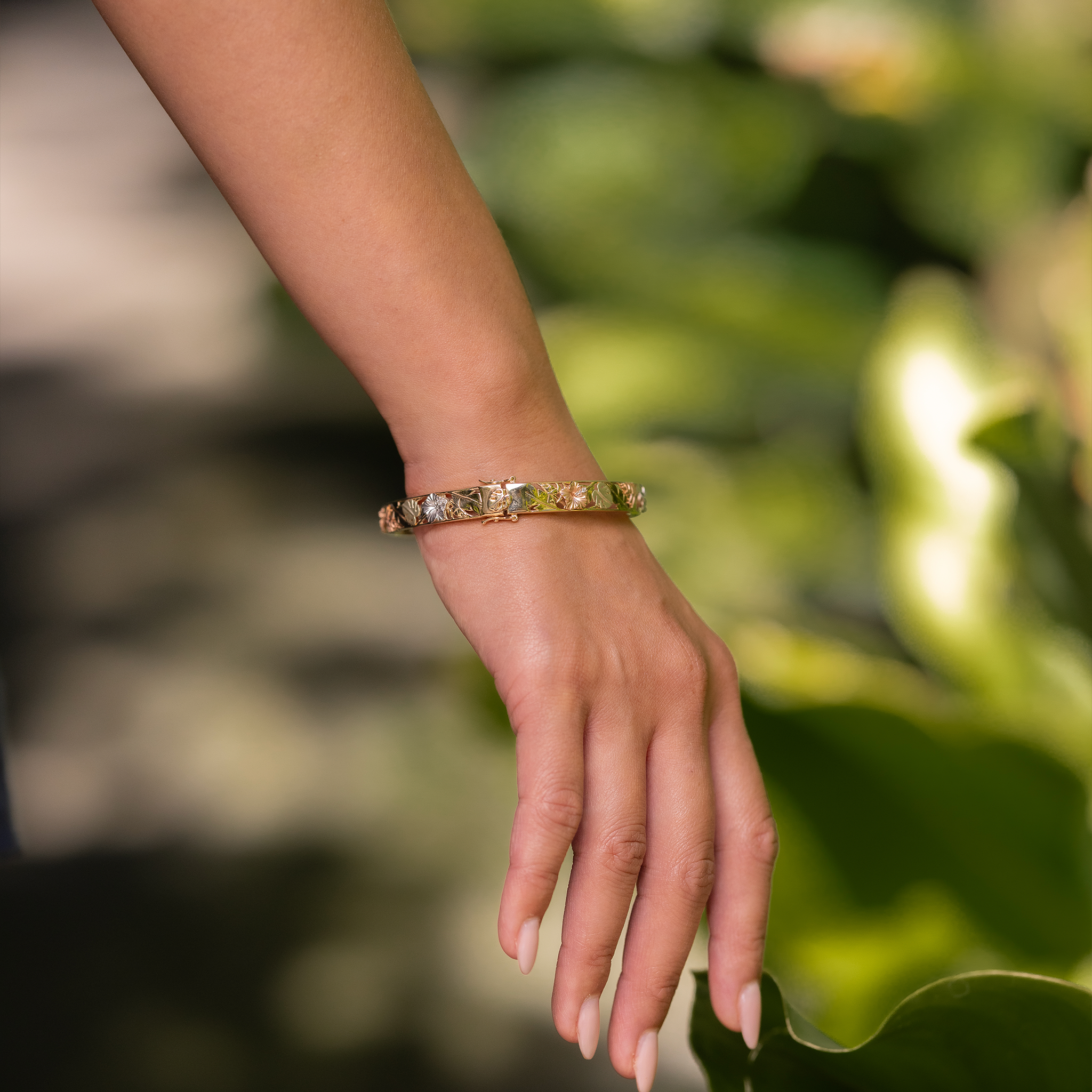 Hand wearing a Hawaiian Gardens Hinge Bracelet in Four Color Gold with Diamonds against a blurred green background