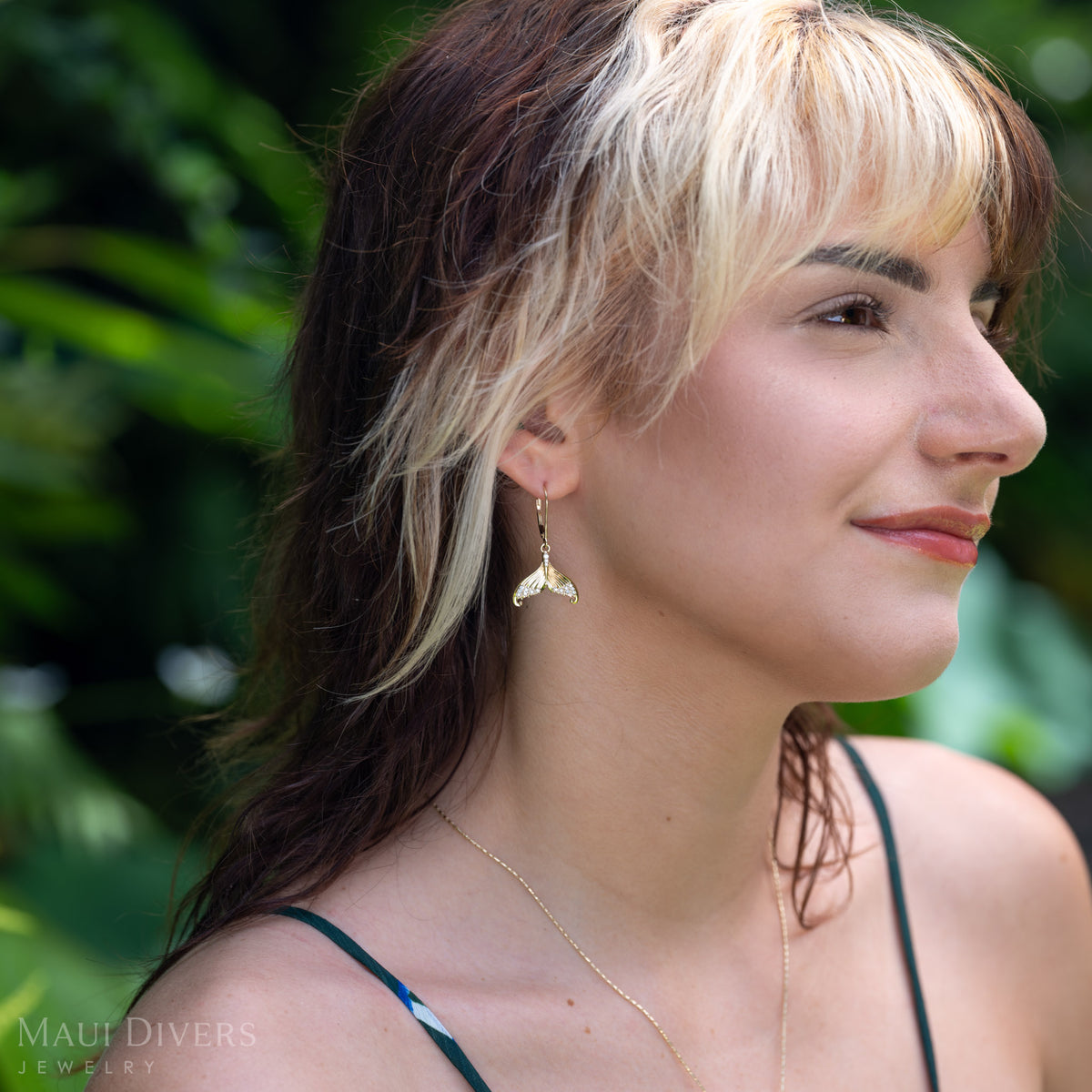A woman in profile wearing a dangling diamond mermaid tail leverback earring in 14k yellow gold outdoors with blurred green foliage in the background