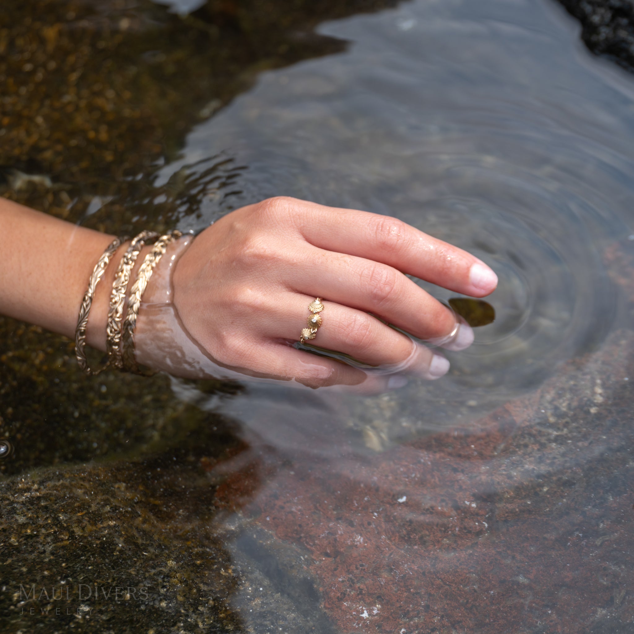 Hand wearing a gold and diamond ʻopihi ring and bracelets touching water