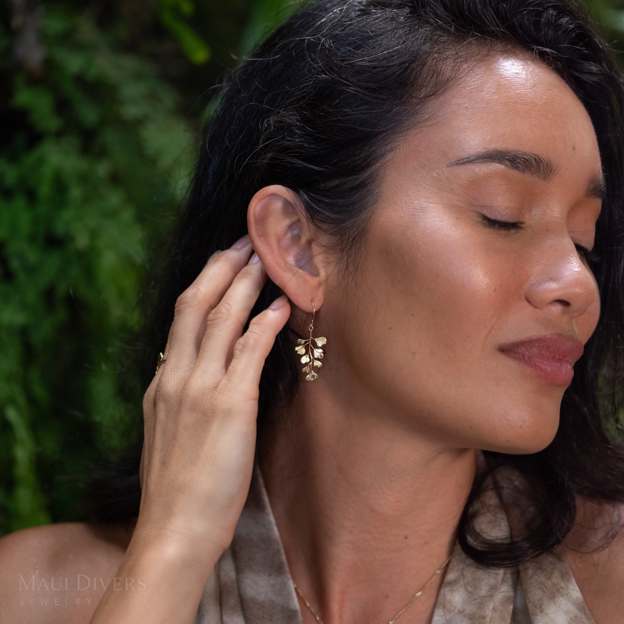 A close-up lifestyle shot of a woman with dark hair wearing a delicate gold dangle maiden hair fern earring, set against a soft-focus green background.
