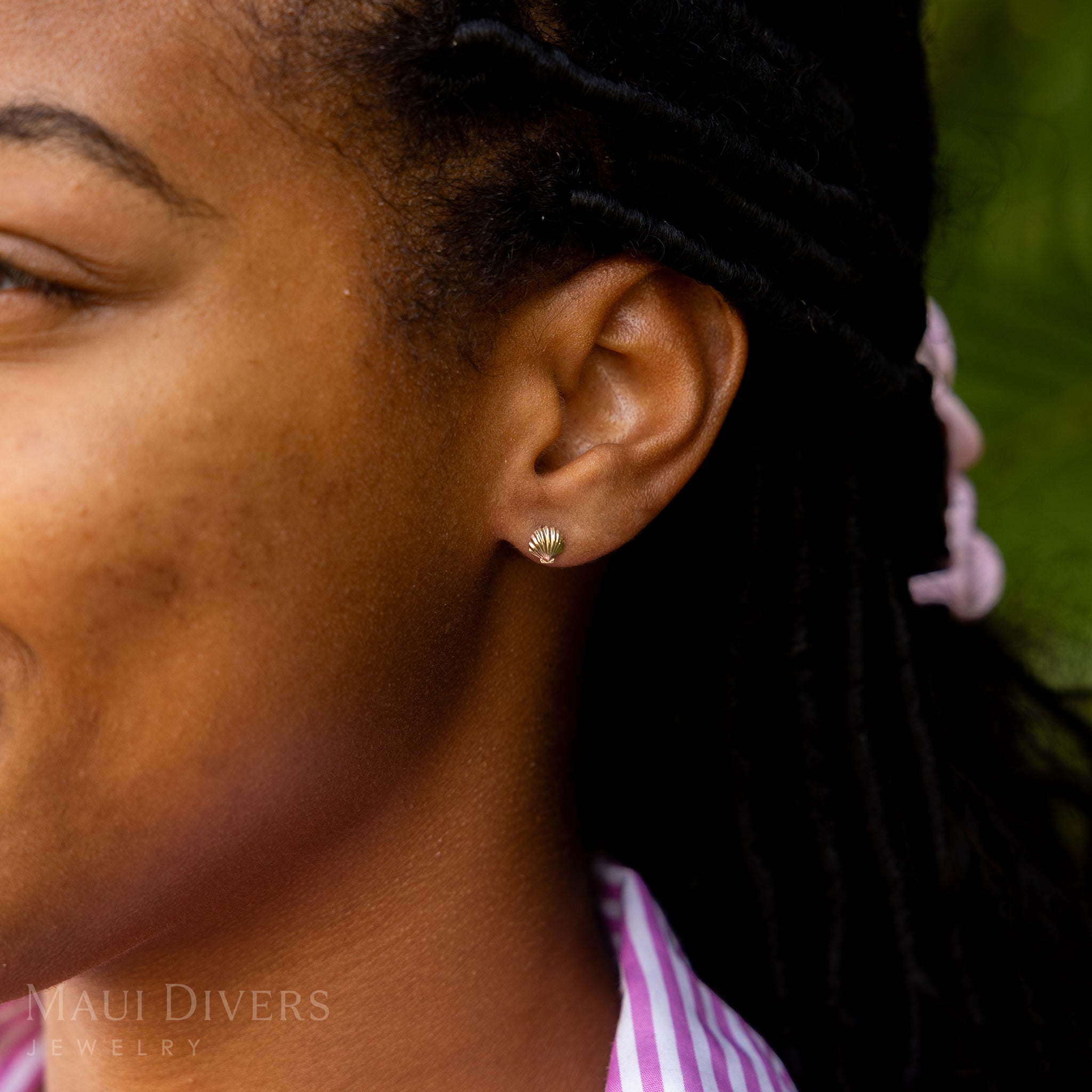A side view close-up of a smiling woman in a pink and white stripe top wearing a Hawaiian Sunrise Shell Earring in 14k yellow gold, against a blurred leafy green background