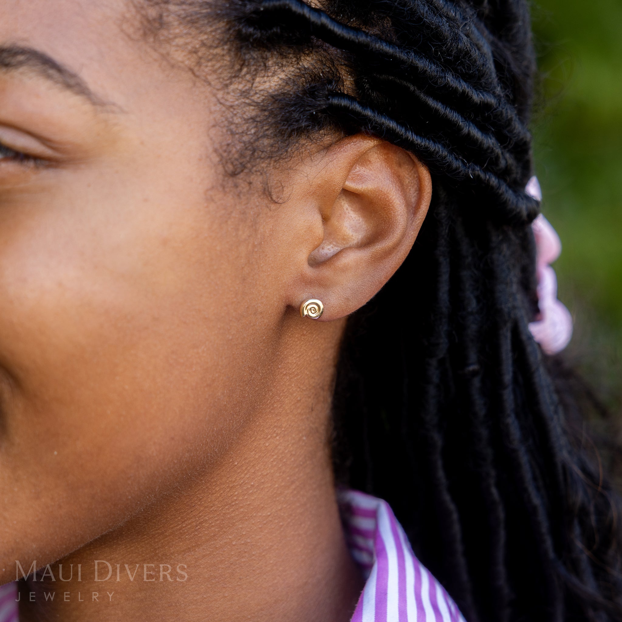 A side view close-up of a woman in a pink and white stripe top wearing a Conch Shell Earring in 14k yellow gold, against a blurred green background