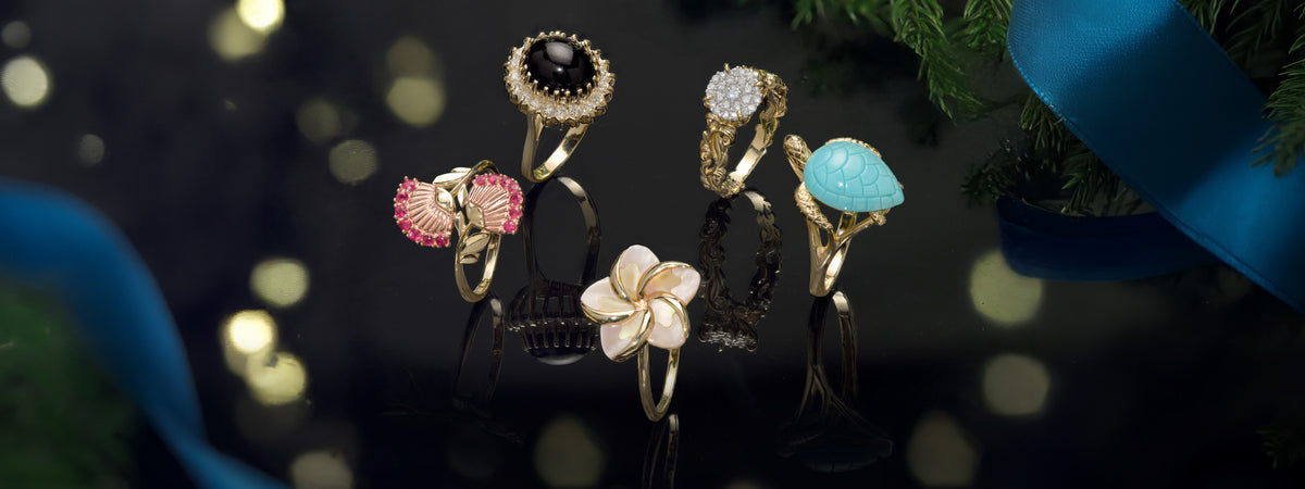 Five yellow gold rings with colorful gemstones in varying designs displaying on a reflective black surface with festive bokeh lights and greenery