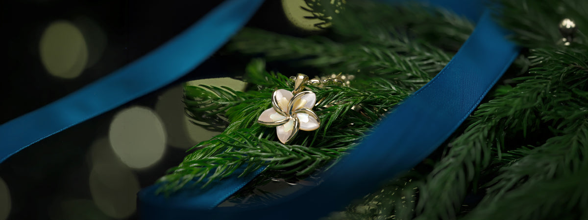 A 14k yellow gold plumeria pendant with iridescent mother-of-pearl petals, resting on green pine branches next to a blue ribbon against a dark, festive background.