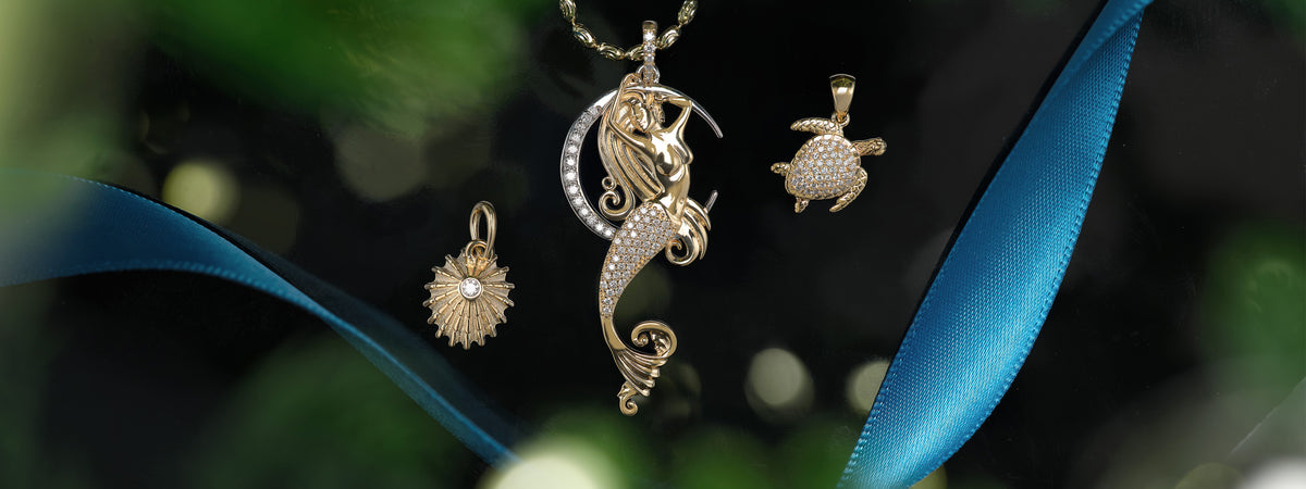 A close-up of several gold pendants against a dark, blurred background with green bokeh and a bright blue ribbon. The central pendant is a mermaid with a crescent moon, flanked by a turtle and 'opihi pendants.