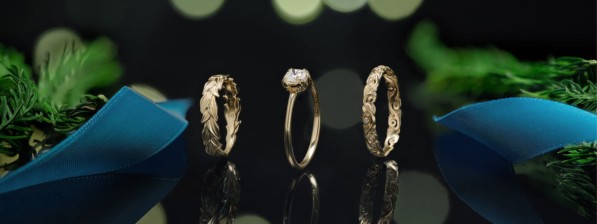 Three gold rings displayed on a reflective black surface, with two ornate textured rings and one ring featuring a diamond. Blue ribbon and green foliage decorate the sides against a dark background with soft green bokeh lights.
