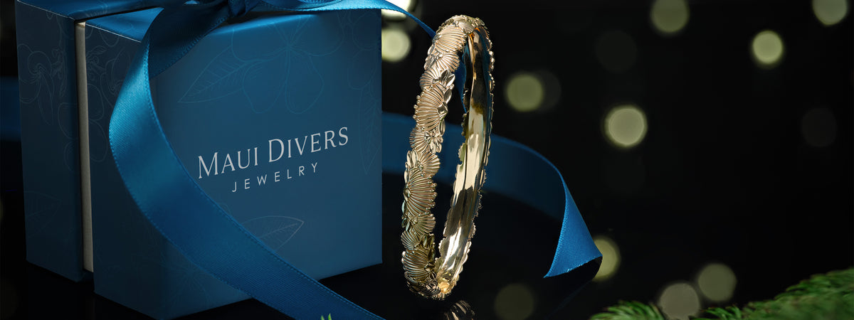 A 14k yellow gold ʻŌhiʻa Lehua Bracelet with a blue Maui Divers Jewelry box and blue ribbon with a pine tree against the side, shown on a black reflective surface