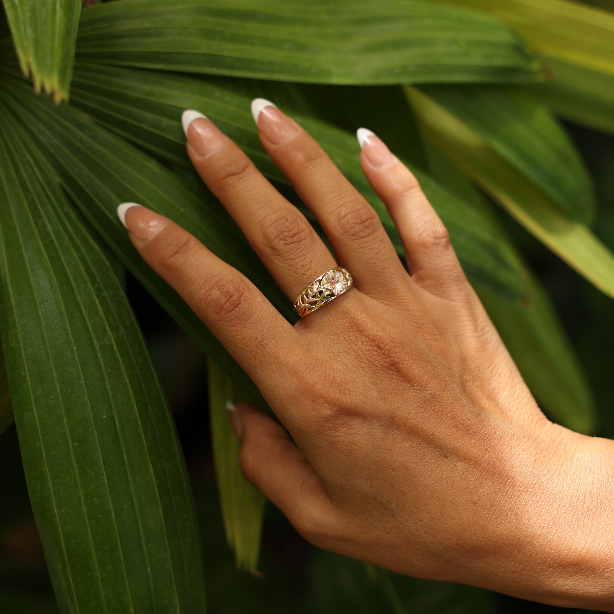 Close up of woman wearing Hawaiian Gardens Hibiscus Ring in Tri Color Gold with Diamonds against green leaves