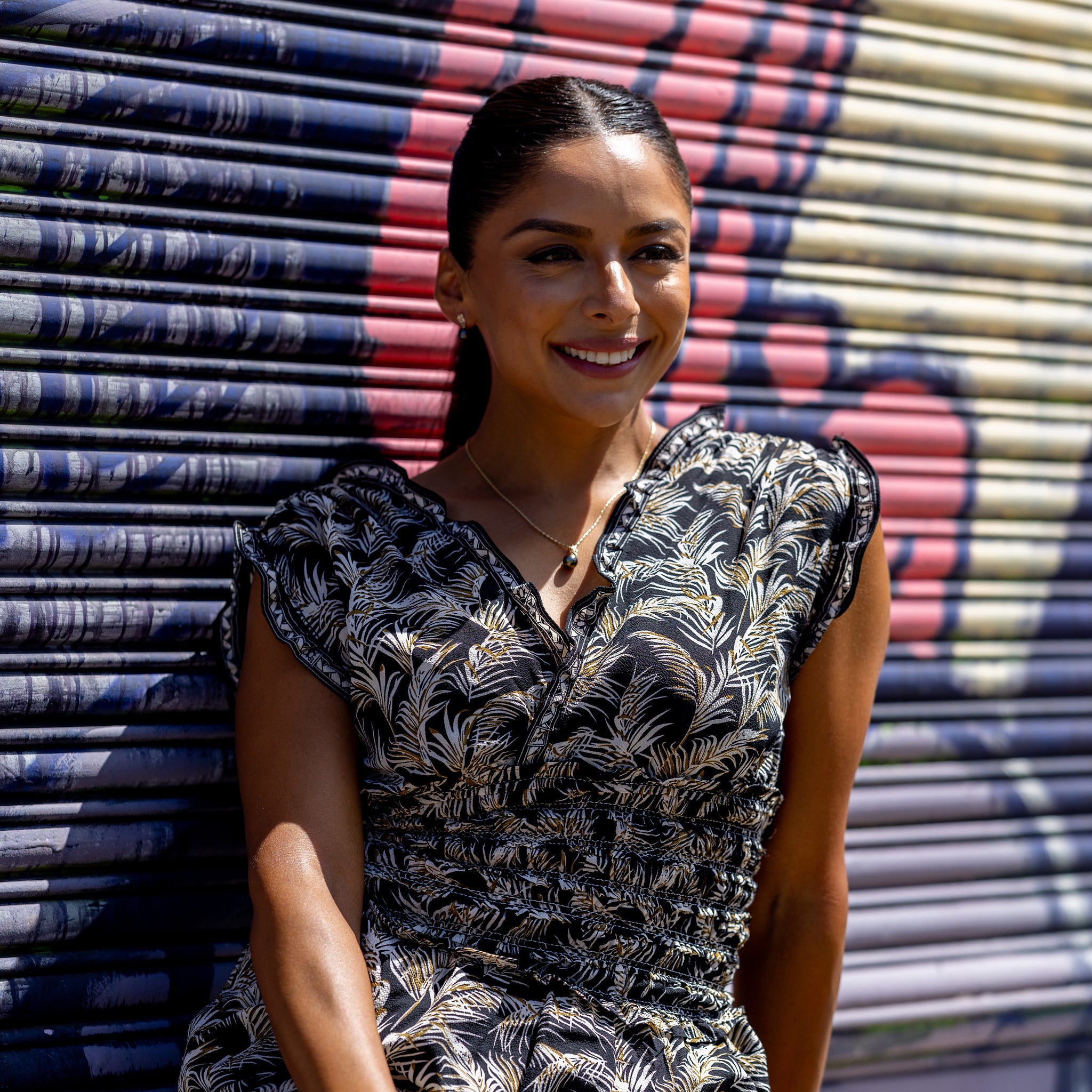 A woman in a black patterned dress wearing a Tahitian black pearl pendant and earrings against a mural background.