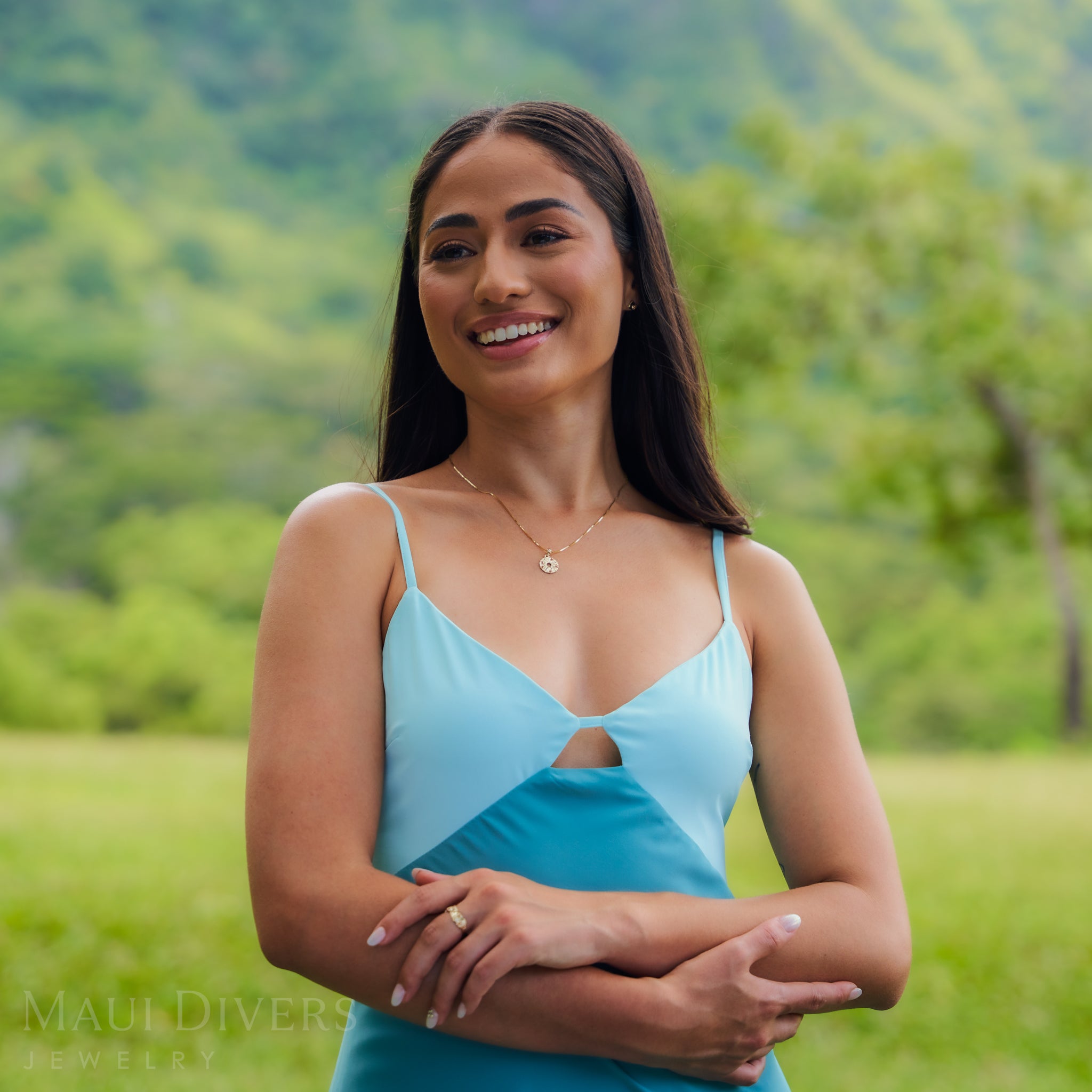 A woman wearing a light blue top and a gold pineapple slice pendant on a chain outdoors.