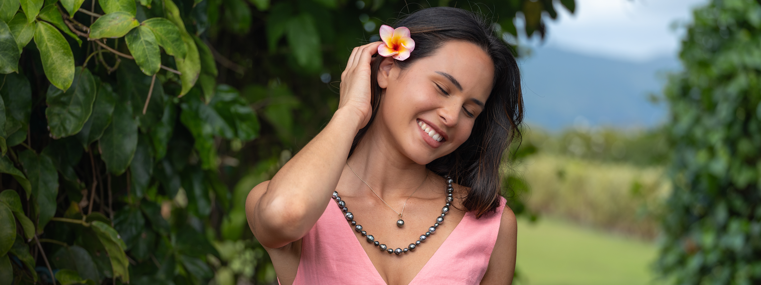 Smiling woman in a pink dress with a plumeria in her hair wearing a Tahitian black pearl strand and Tahitian black pearl pendant in 14k yellow gold with a scenic garden background