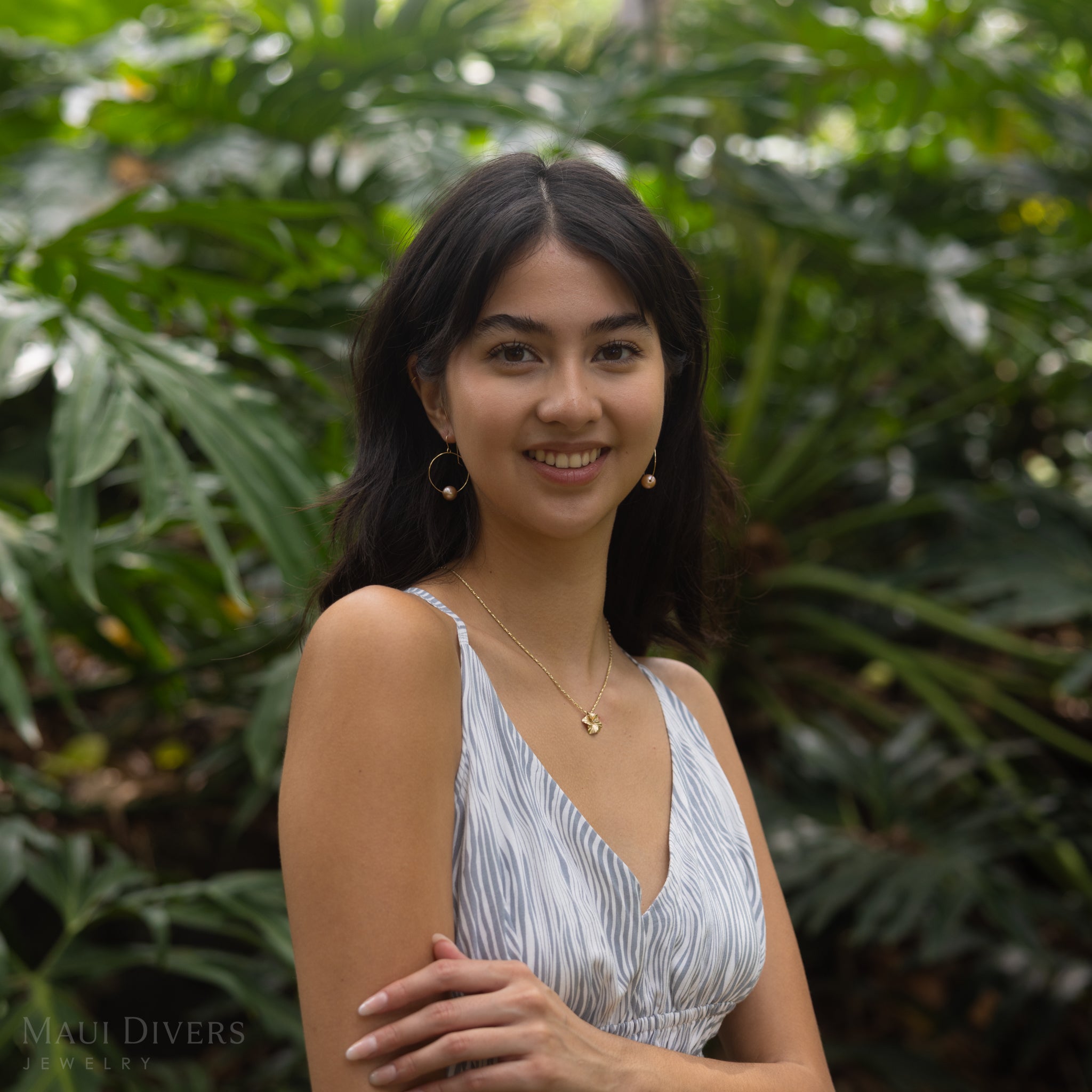 Smiling woman wearing pearl hoop earrings and a diamond hibiscus pendant with a blurry monstera leaf background