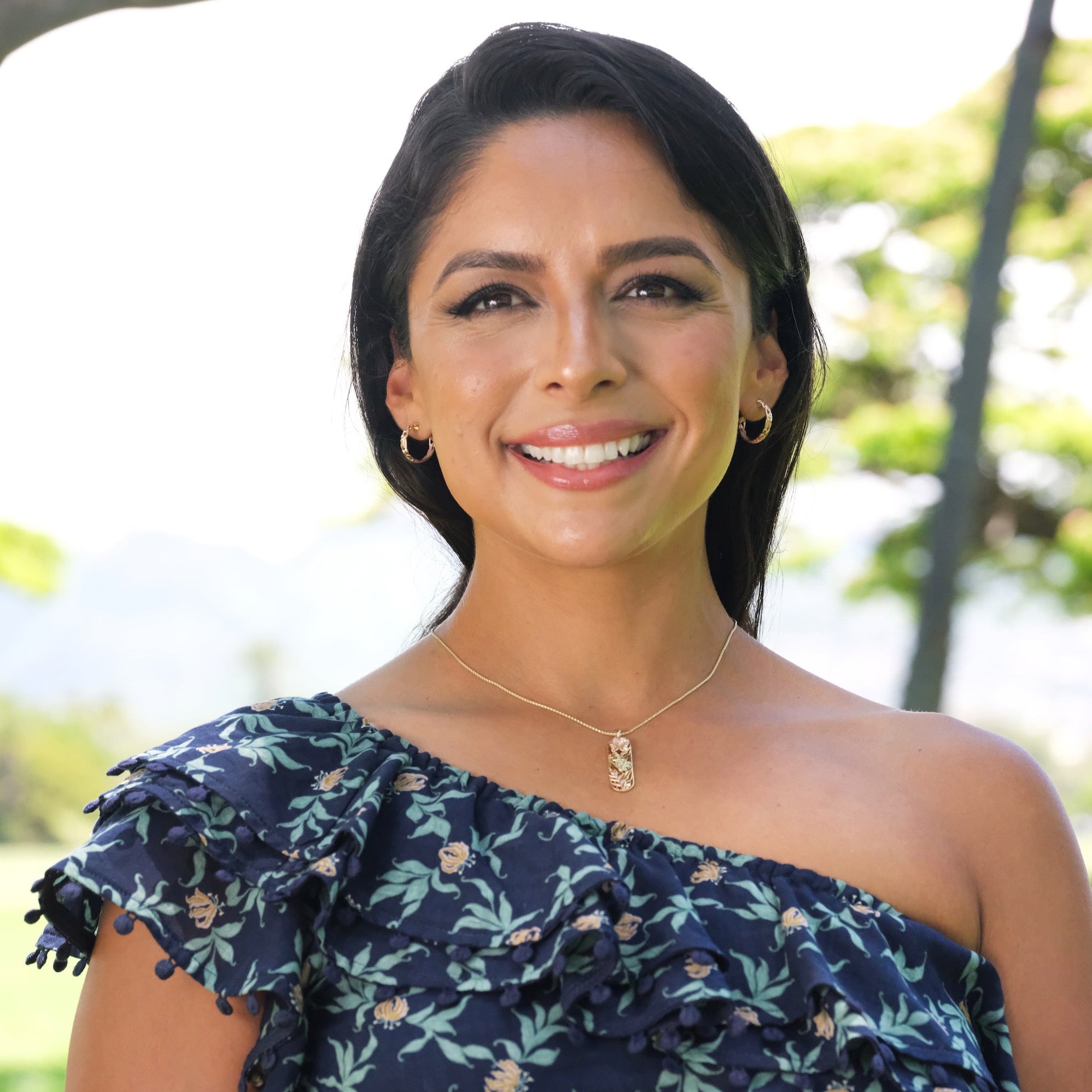 A woman wearing a Hawaiian Gardens Hibiscus Pendant in Tri Color Gold with Diamonds and gold hoop earrings