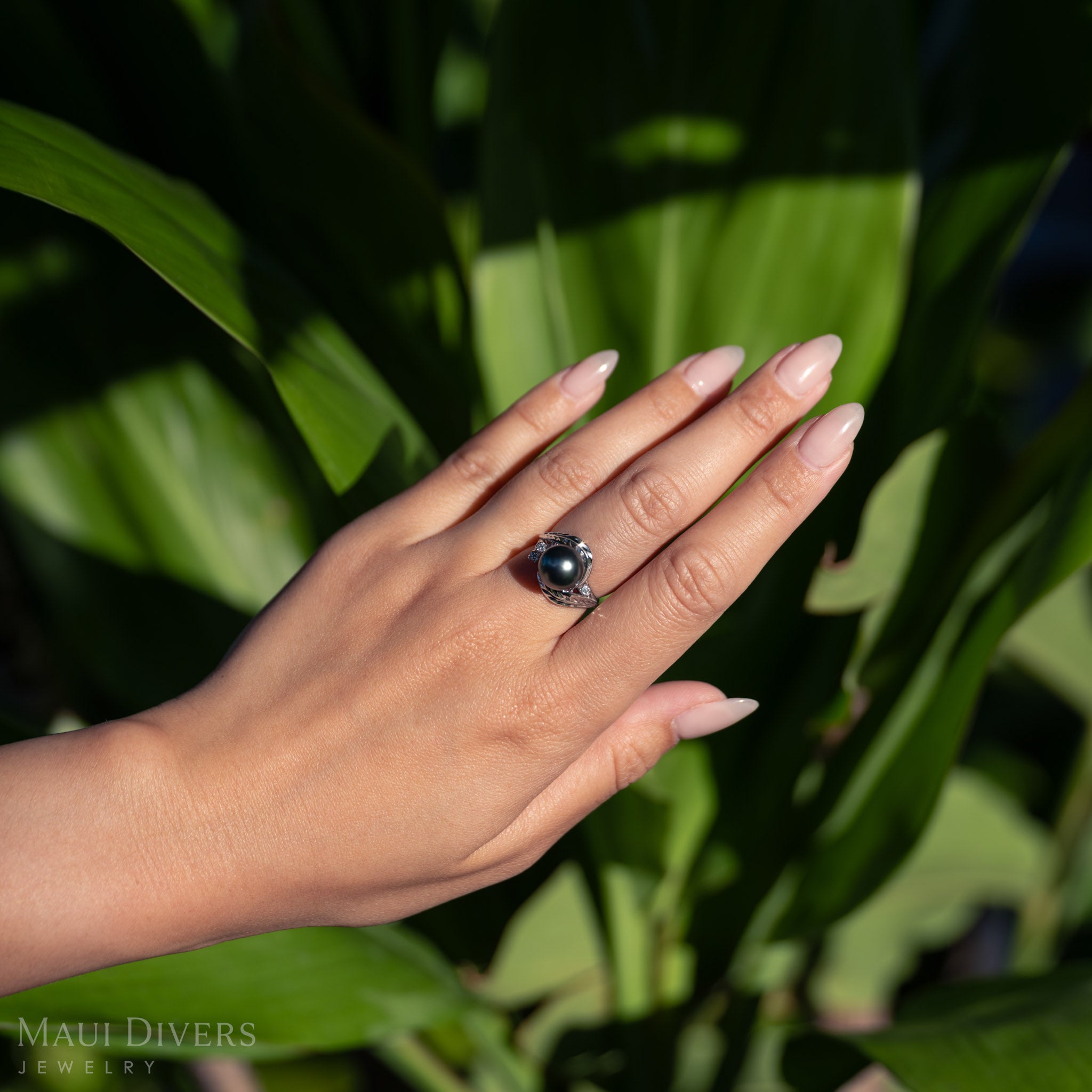 Hand wearing a Maile Leaf Tahitian black pearl ring against a green leafy background