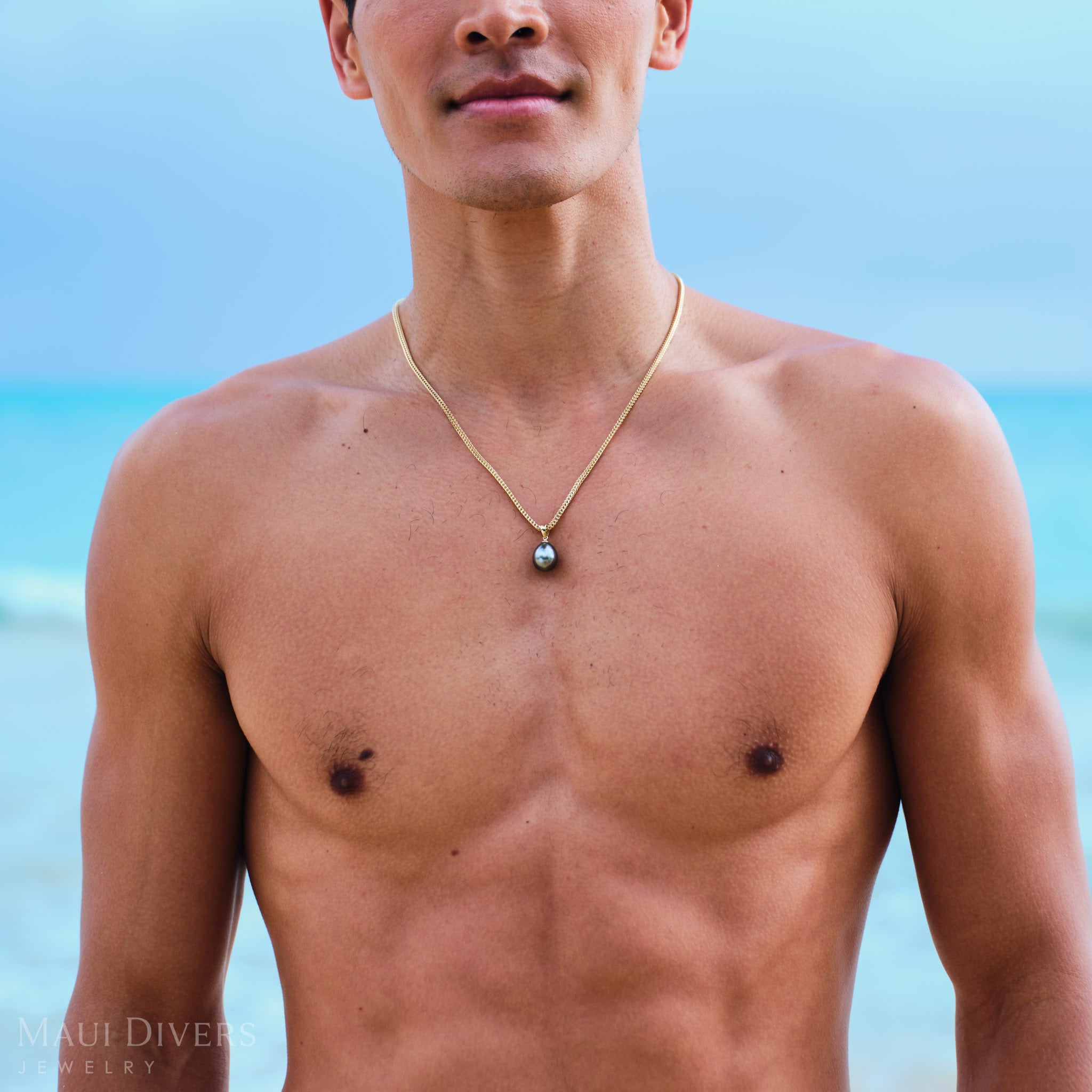 Man wearing a gold necklace with a Tahitian black pearl pendant pendant on a beach