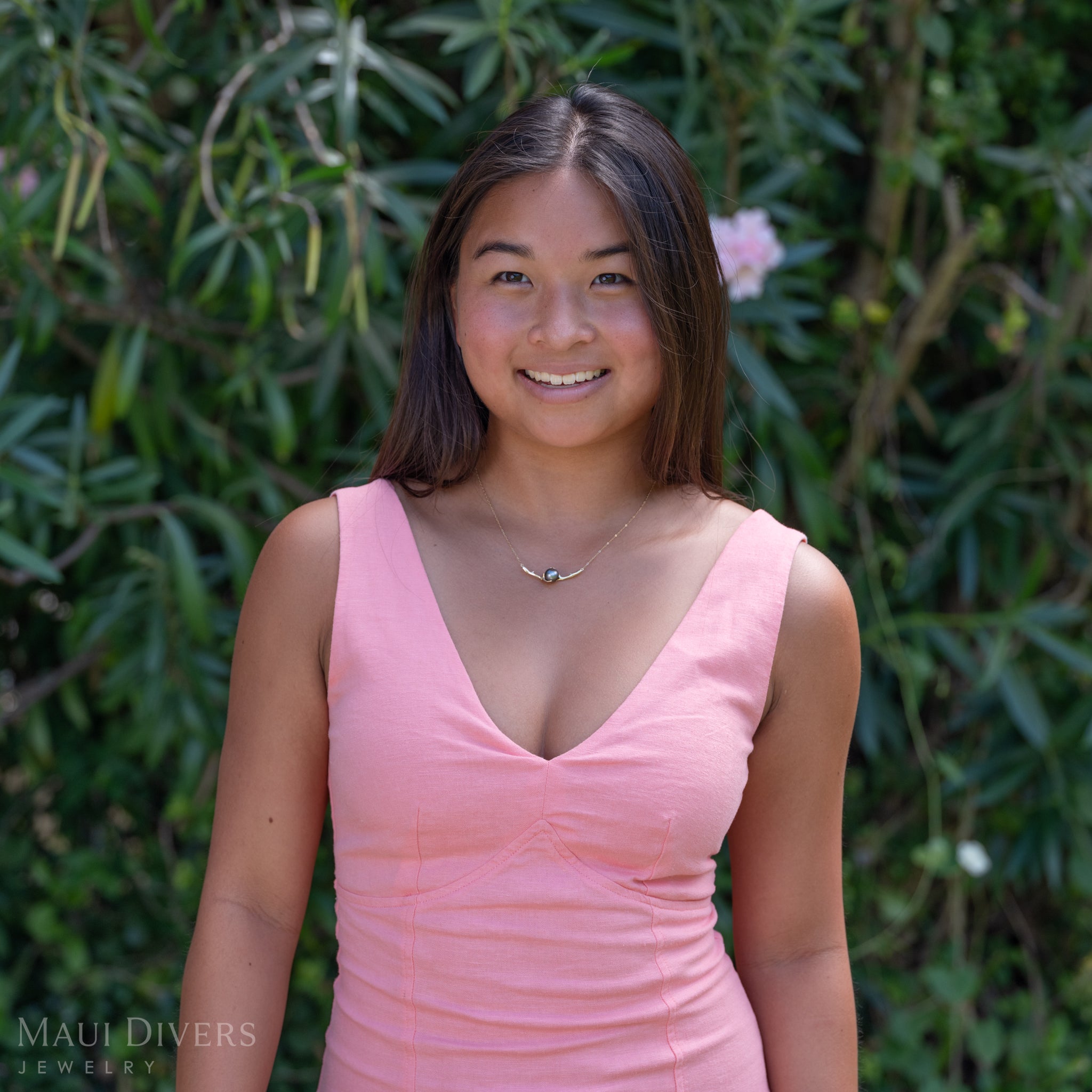 A smiling woman in a light pink top wearing a Heritage Tahitian Black Pearl Necklace in 14k yellow gold against a blurred leafy background