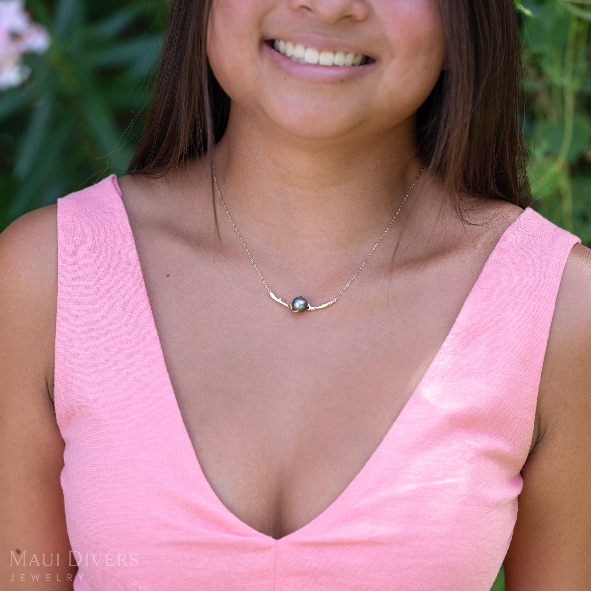 A close-up of a smiling woman in a light pink top wearing a Heritage Tahitian Black Pearl Necklace in 14k yellow gold against a blurred leafy background