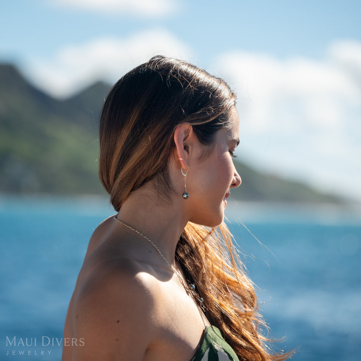 A woman wearing a gold Heritage Collection Leverback Earring with a Tahitian black pearl against a blurry mountain and ocean background