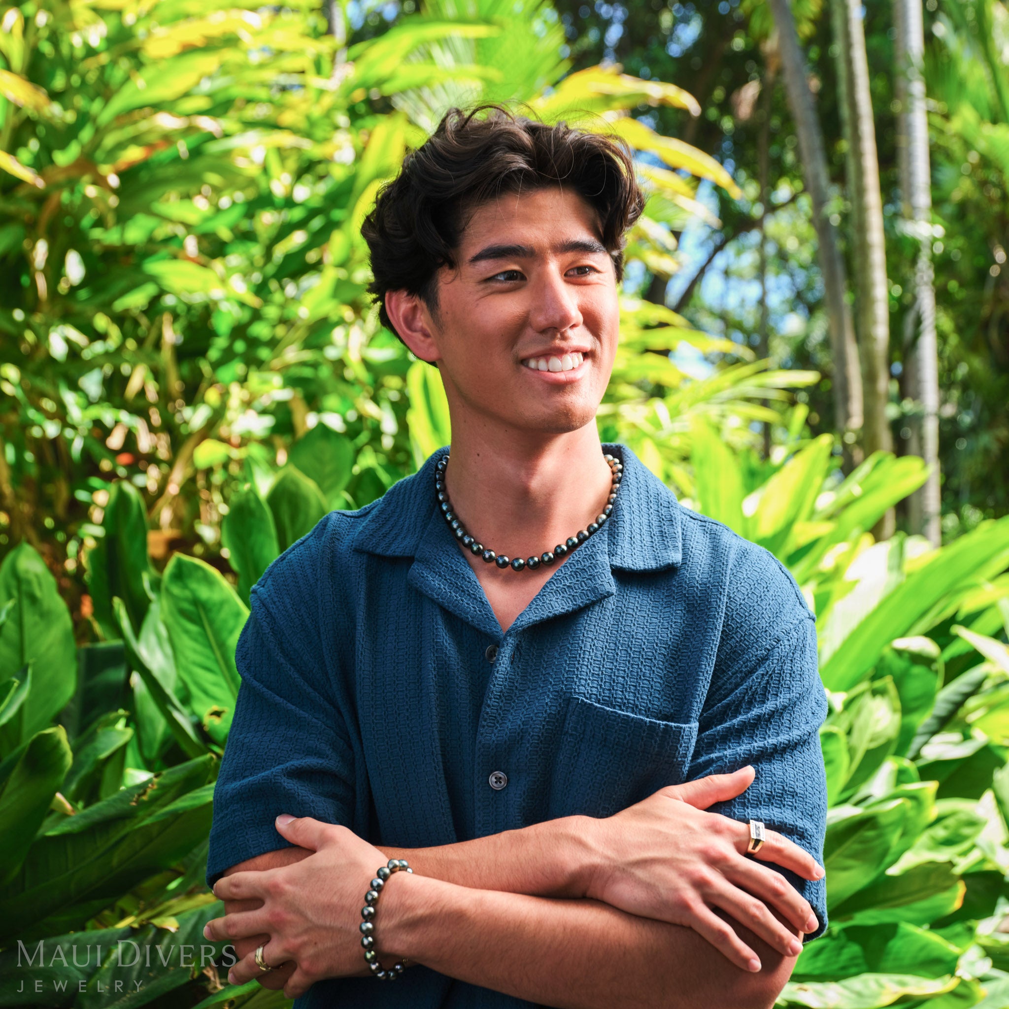 Smiling man in a blue shirt wearing a Tahitian black pearl strand necklace with a matching Tahitian black pearl bracelet, against a blurred forest background