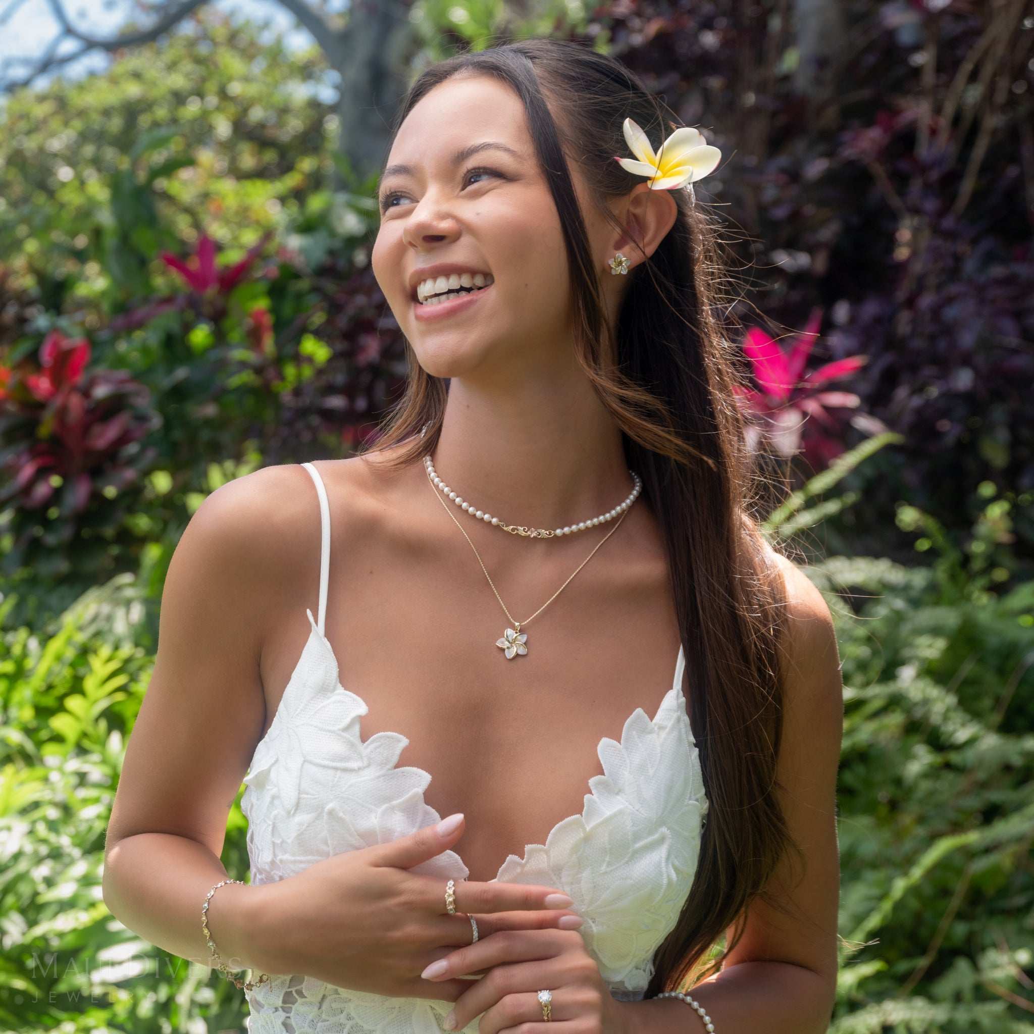 A woman outdoors wearing a Plumeria mother-of-pearl pendant on a 14k yellow gold chain, including a pearl choker with a Hawaiian Heirloom plumeria design in 14k rose gold yellow gold, along with rings and a bracelet. She is smiling and looking to the side, wearing a white ruffled top with a white plumeria flower tucked behind her ear, set against a lush green garden background.