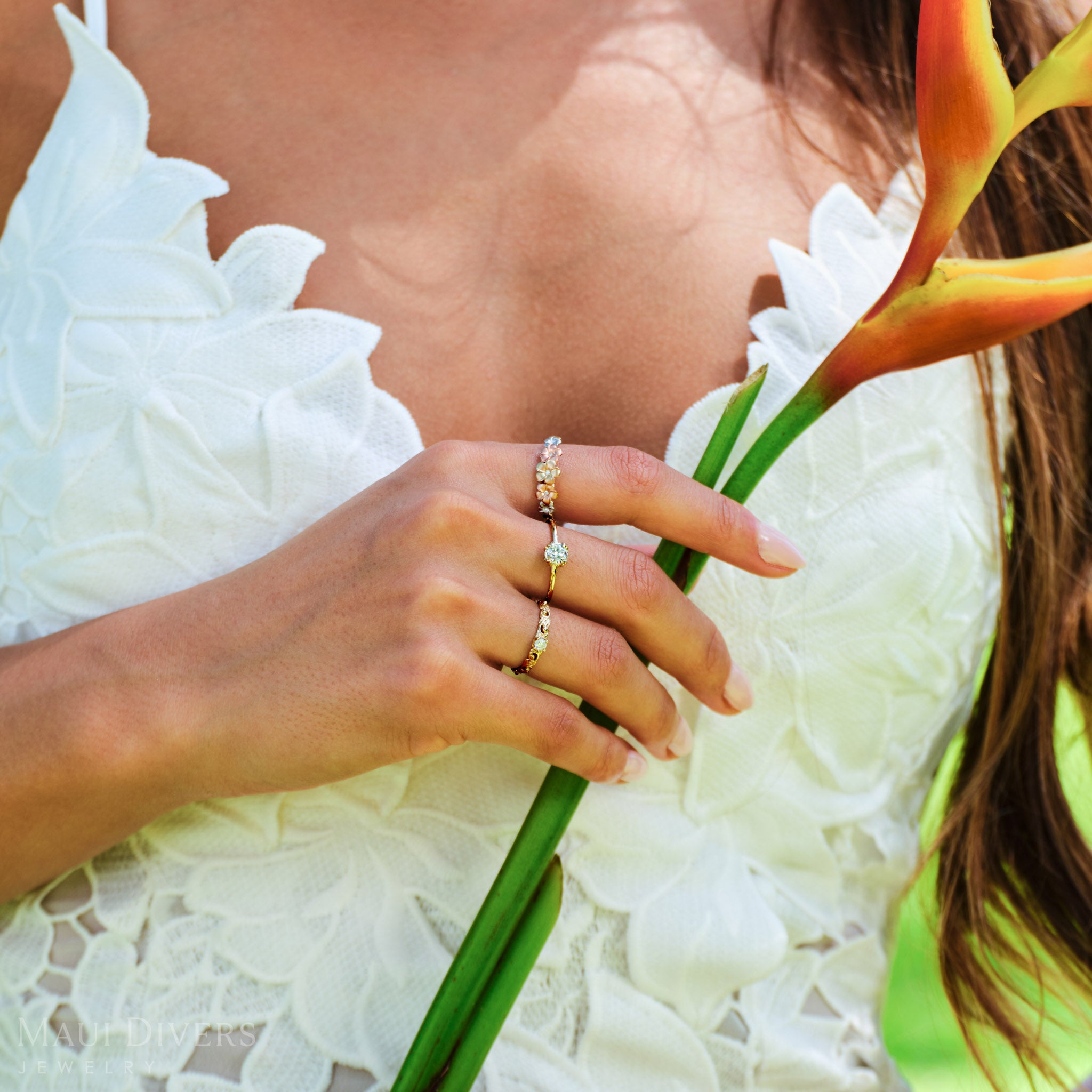 Close-up of a hand wearing a Plumeria Ring in Tri Color Gold with Diamonds, Hidden Hawaiʻi Maile Ring in Gold with Lab Grown Diamond, and a Living Heirloom Ring in 14k Yellow Gold with Diamond holding a Heliconia flower stem 