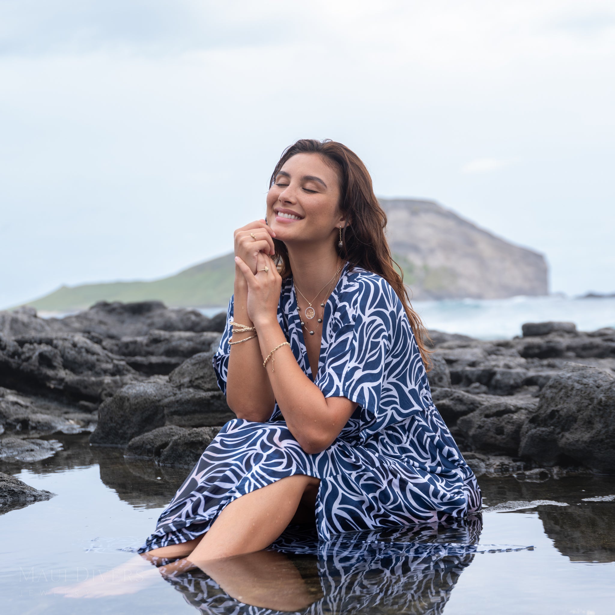 Woman wearing a blue and white patterned top with a five pearl Tahitian black pearl necklace and a diamond coral branch design pendant sitting in tid pool along a rocky beach