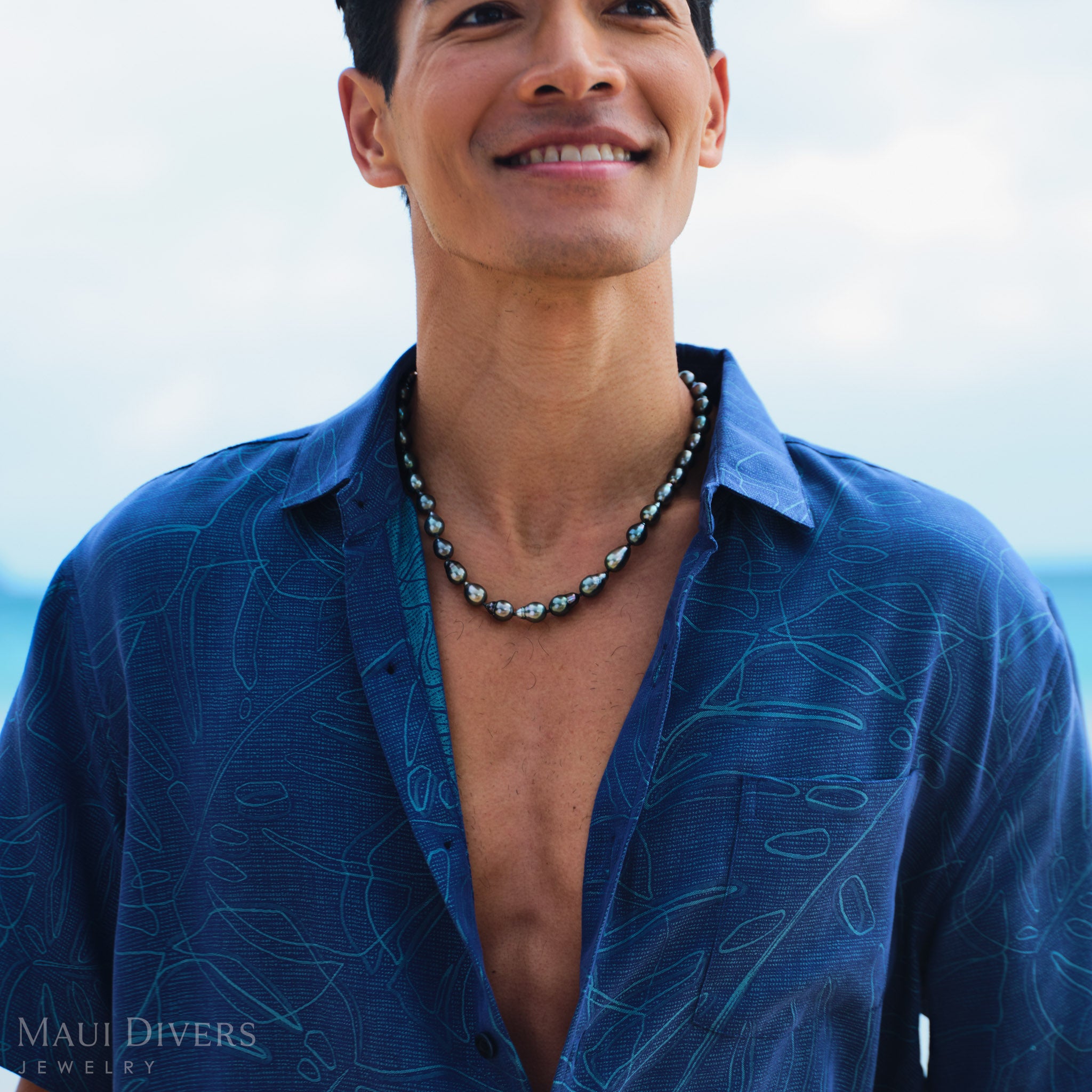 Man wearing a blue patterned shirt and Tahitian black pearl strand with a blurred background