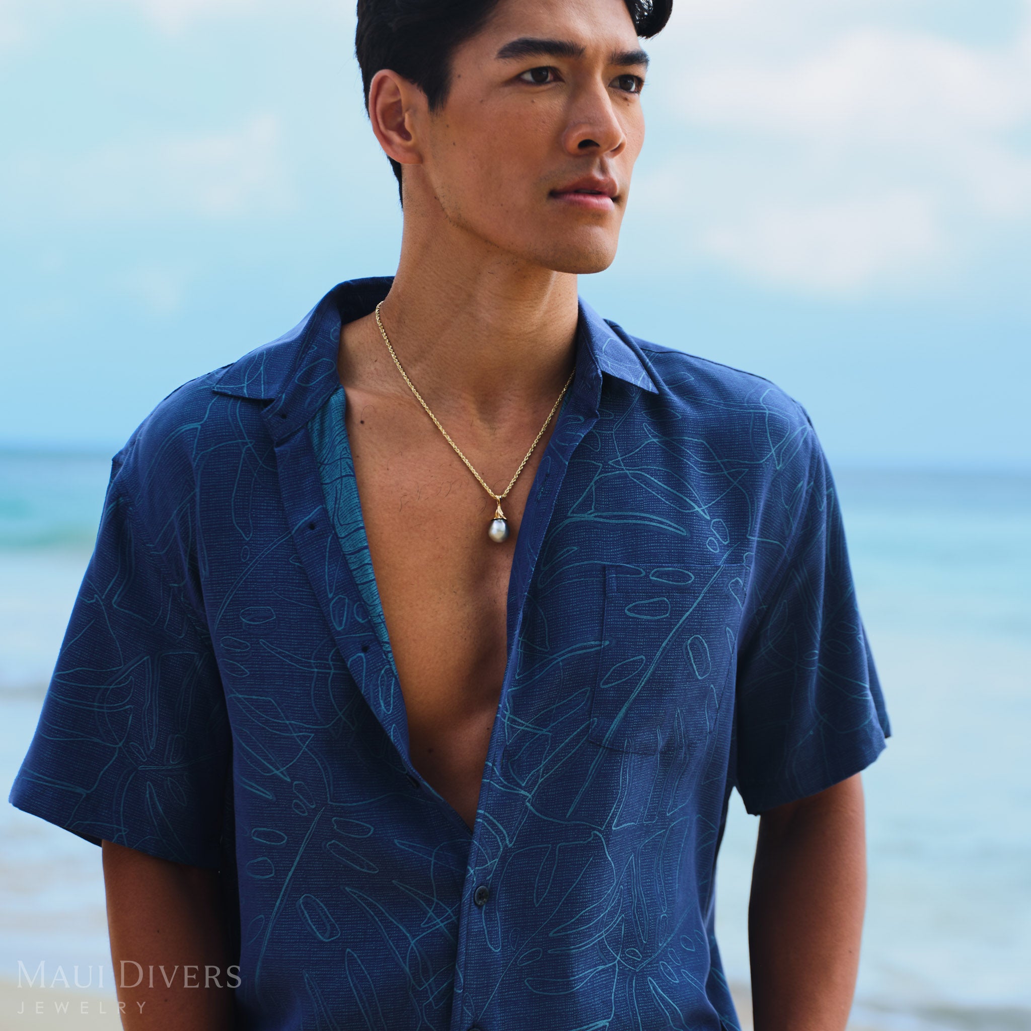 Man wearing a blue patterned shirt with a gold waterfall Tahitian black pearl pendant, set against a blurred beach background.
