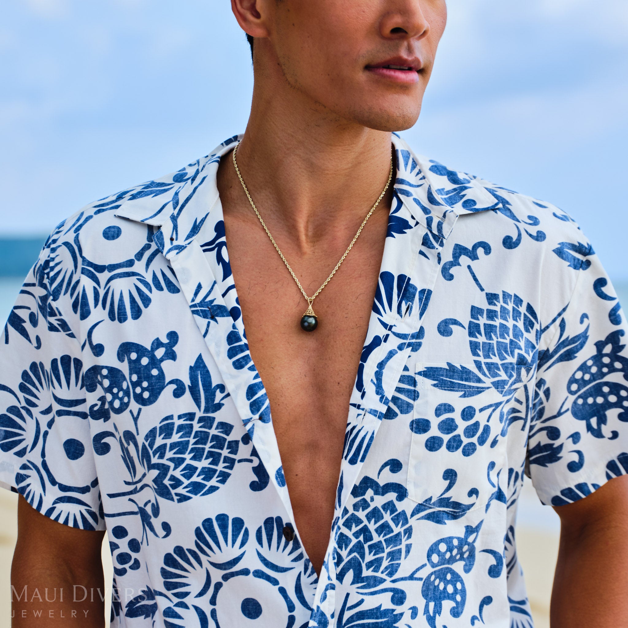 Man wearing a blue and white patterned shirt with a gold fish net Tahitian black pearl necklace, standing against a clear sky.