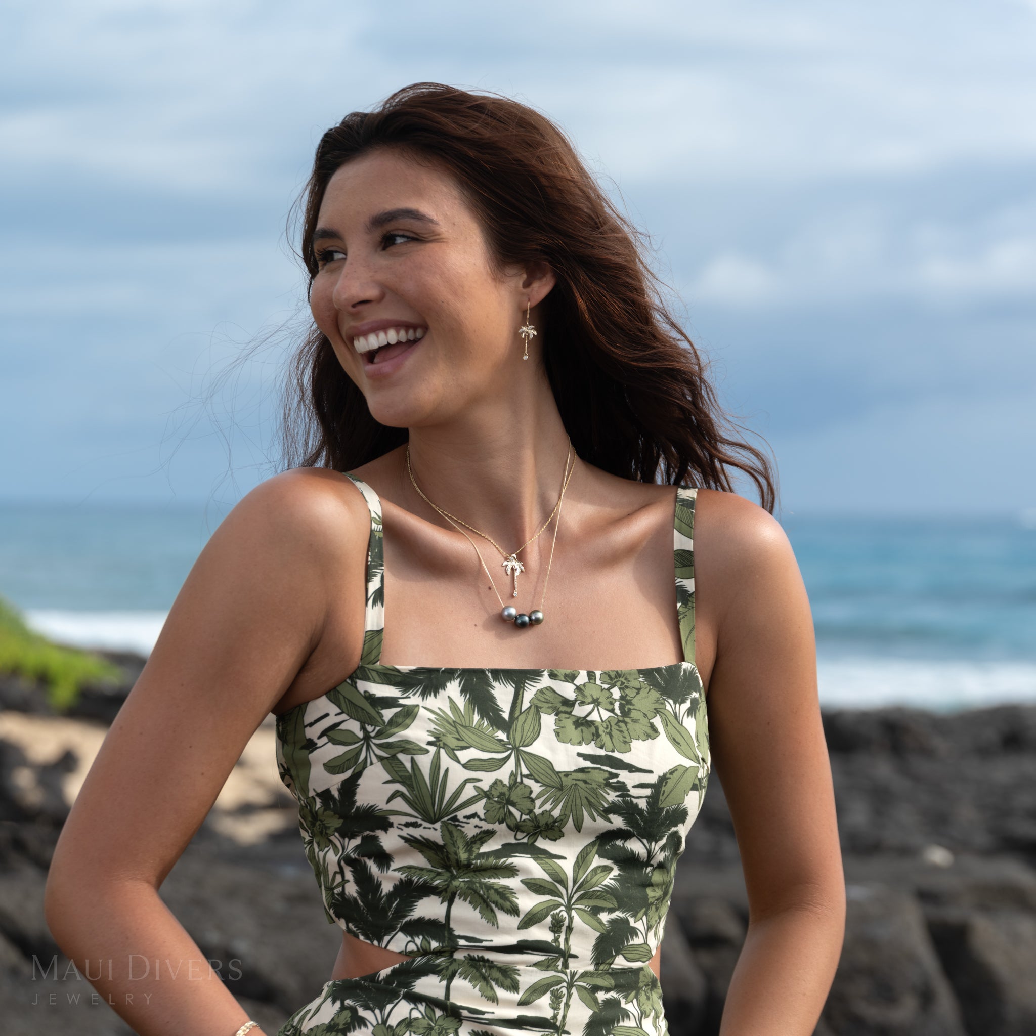 Woman wearing a green-and-white patterned top, gold and diamond palm tree earrings, a gold necklace featuring three varying-color Tahitian black pearls, and a layered diamond palm tree pendant on a rope chain, standing on a rocky beach with an ocean view