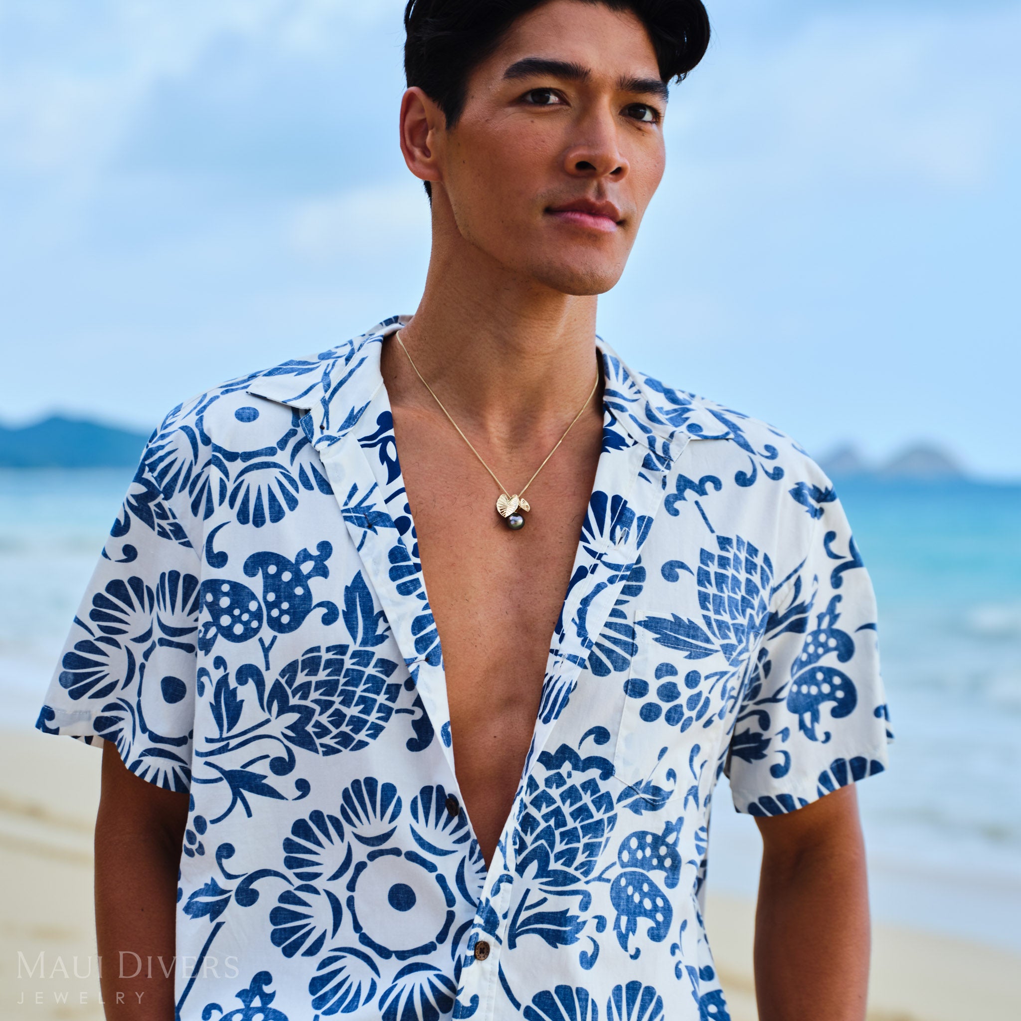 Man wearing a blue and white patterned shirt with a Kalo Tahitian black pearl necklace, on a beach.