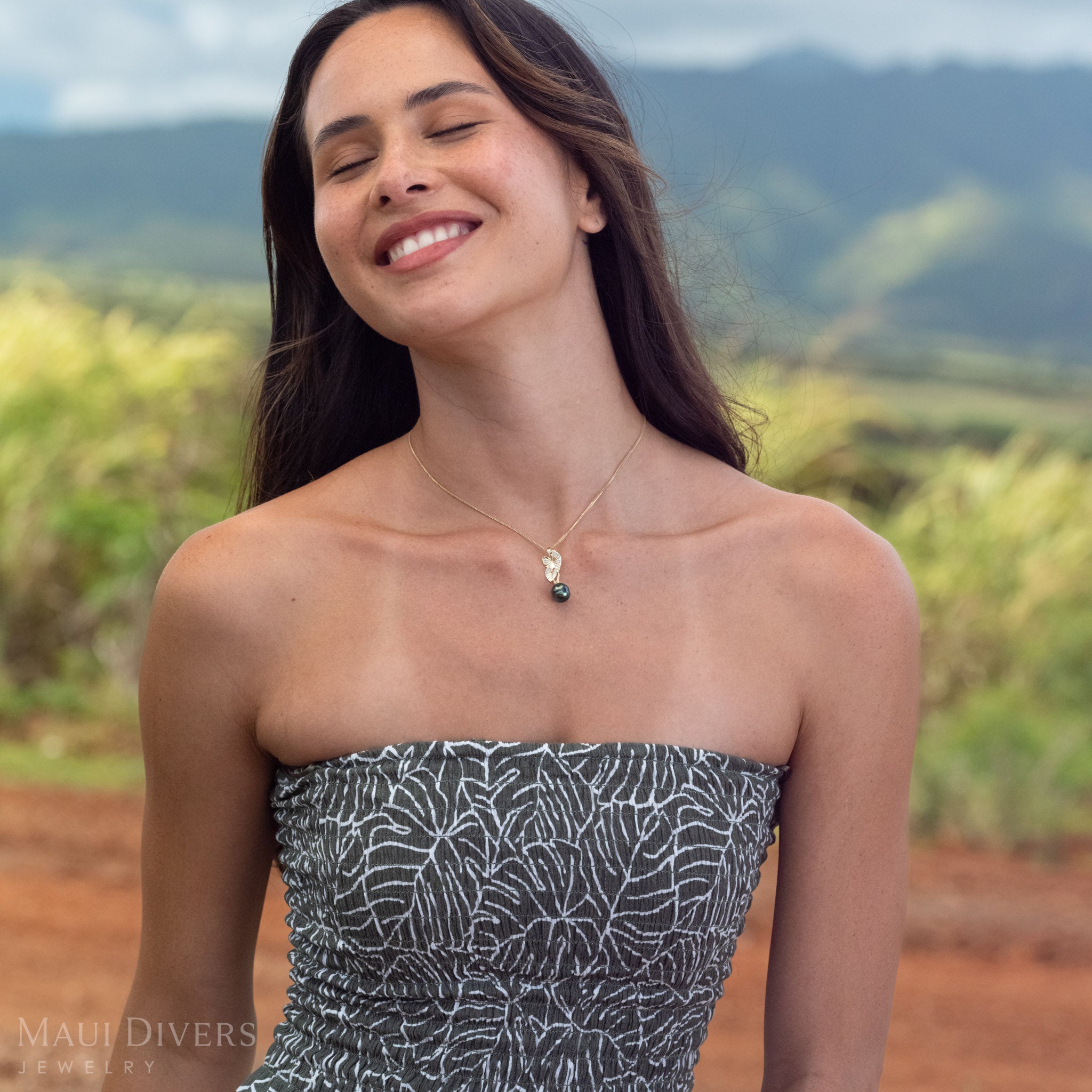 A woman smiling wearing a 14k Gold Kalo (Taro) Pendent with a Black Tahitian Pearl in front of a blurred green mountain background. 