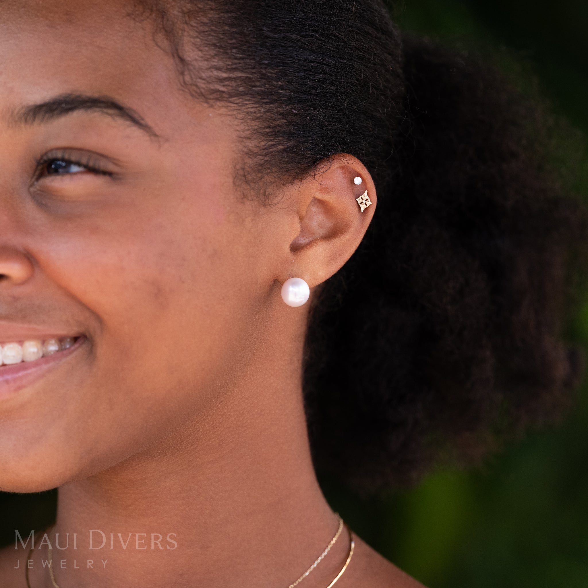 A close-up of a smiling woman wearing a Freshwater White Pearl Stud Earrings in 14k yellow gold, a Hawaiian quilted star stud earrings in 14k yellow gold and a diamond stud earring, against a blurred leafy green background