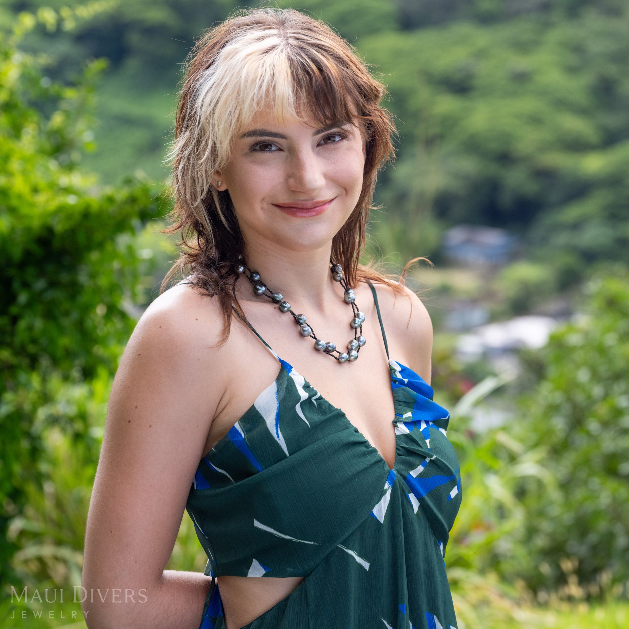 A woman outdoors wearing a dark green halter dress with blue and white abstract patterns, accessorized with a double-layered baroque Tahitian black pearl and black spinel necklace. The background is a soft-focus green landscape.