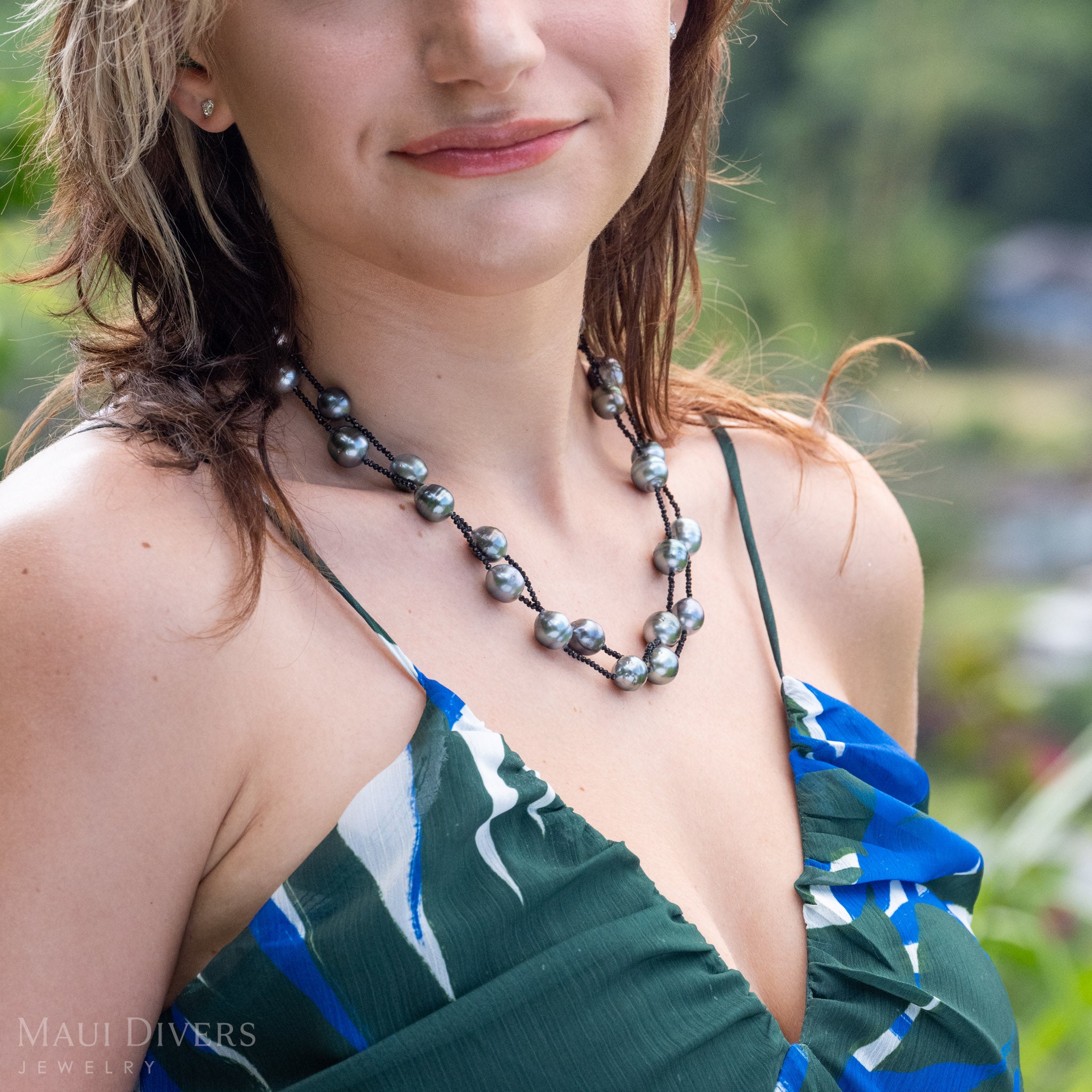 A close-up lifestyle shot of a woman wearing a double-layered baroque Tahitian black pearl and black spinel necklace. She is wearing a green and blue patterned top, with a blurred tropical outdoor background.