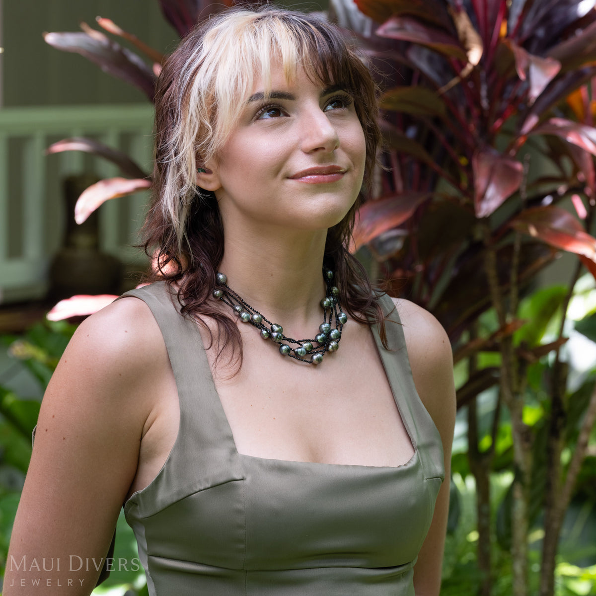A woman wearing a chunky multi-layered Tahitian black pearl and black spinel necklace with dark green and silver beads, styled with an olive green top, set against a tropical garden background with purple-leafed plants.